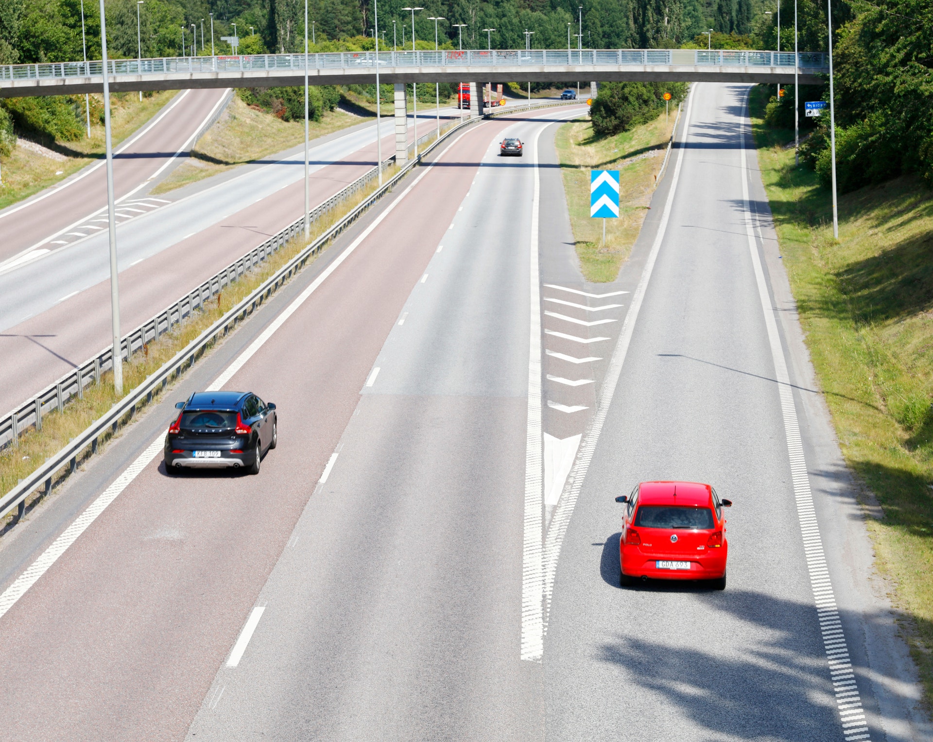 Flygfoto över en motorväg med två röda bilar som kör på olika körfält. En vit bro går över vägen, och gröna träd syns längs vägsidorna.