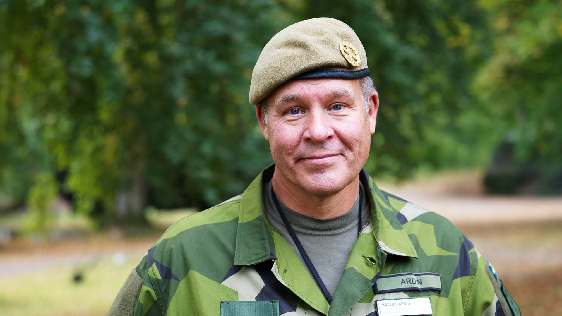 En man i militär uniform och basker ler mot kameran, med skog i bakgrunden.