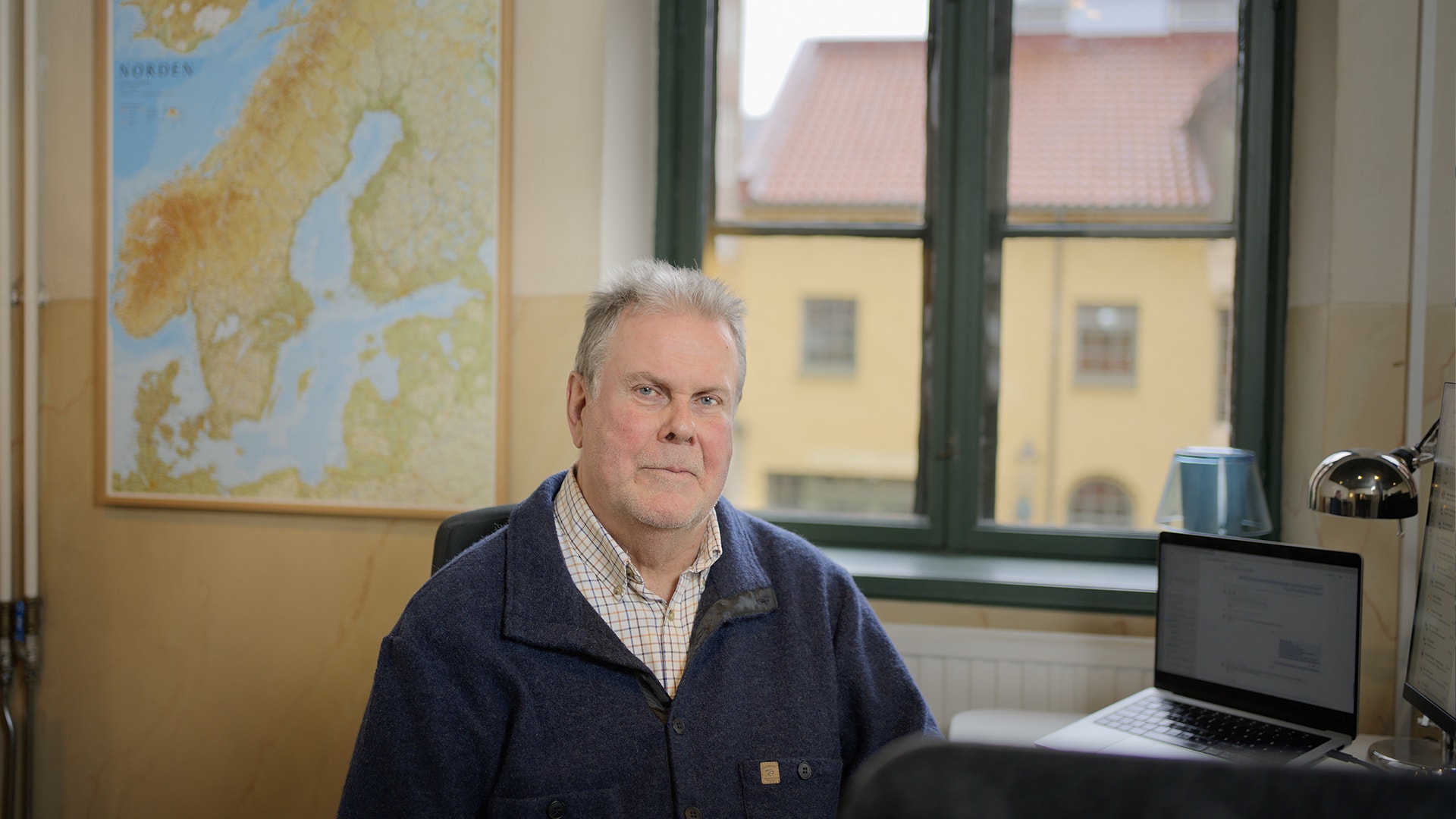 En äldre man sitter vid sitt skrivbord på ett kontor med en dator och en karta på väggen bakom honom.