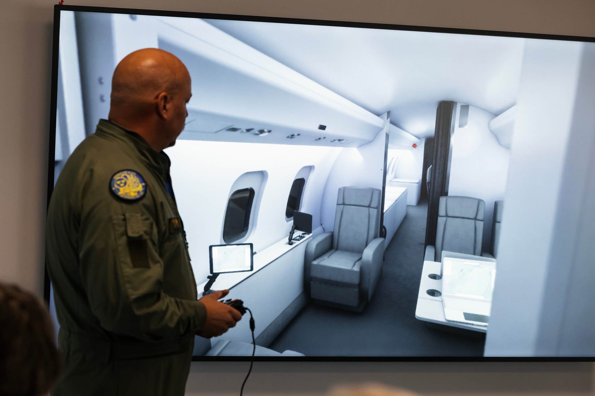 En man i militärgrön uniform står vid en stor skärm som visar en digital bild av en ljus flygplanskabin med fåtöljer.