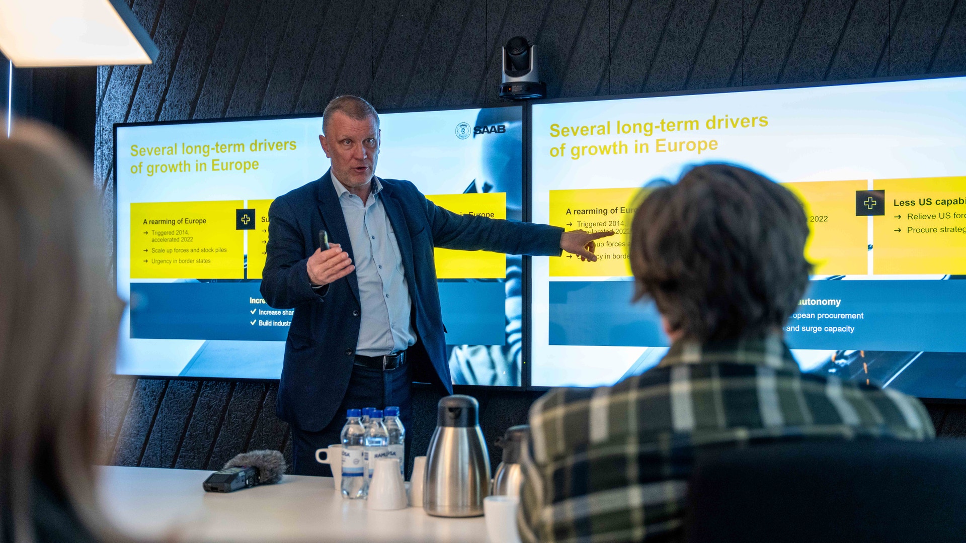 En man håller ett föredrag framför två stora presentationsskärmar med gul och blå design. I förgrunden syns två lyssnare.
