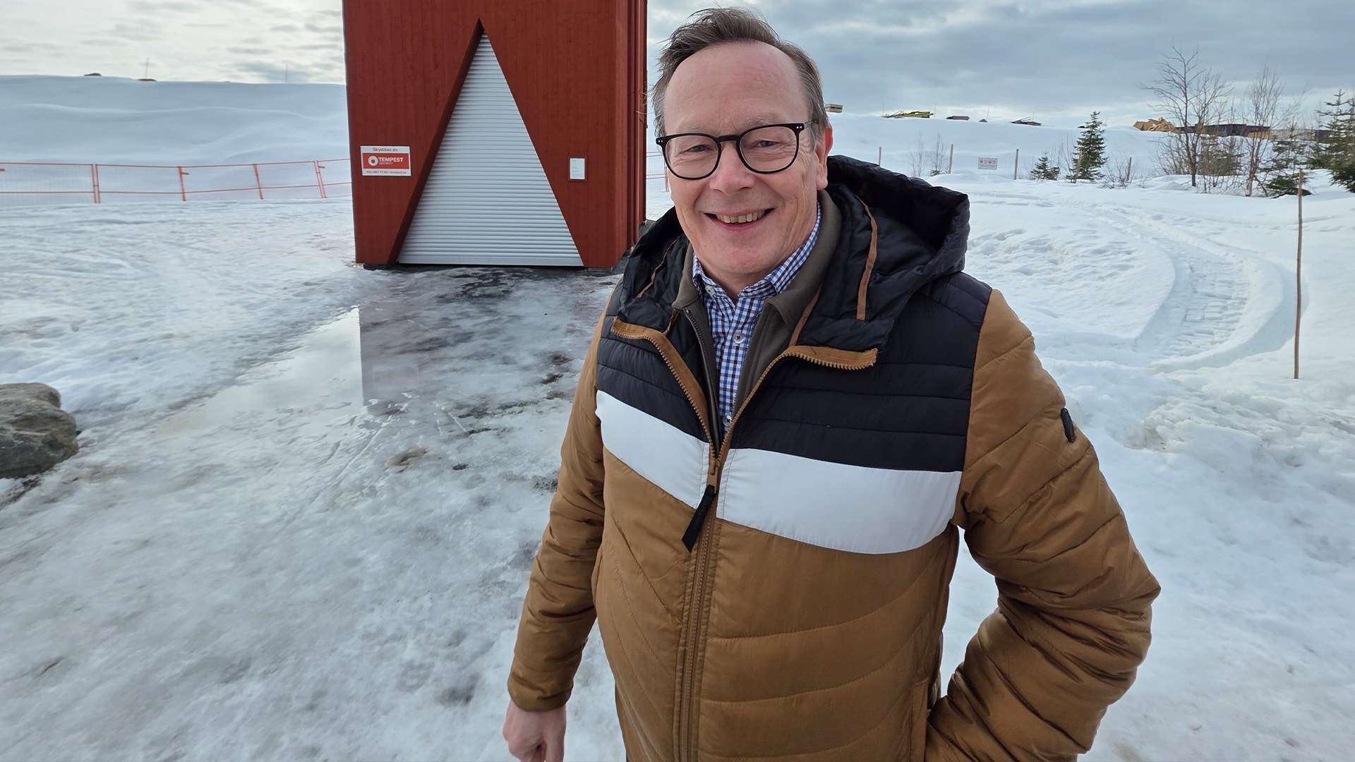 En man i vinterkläder står på en frusen sjö med ett rött isfiskehus i bakgrunden.