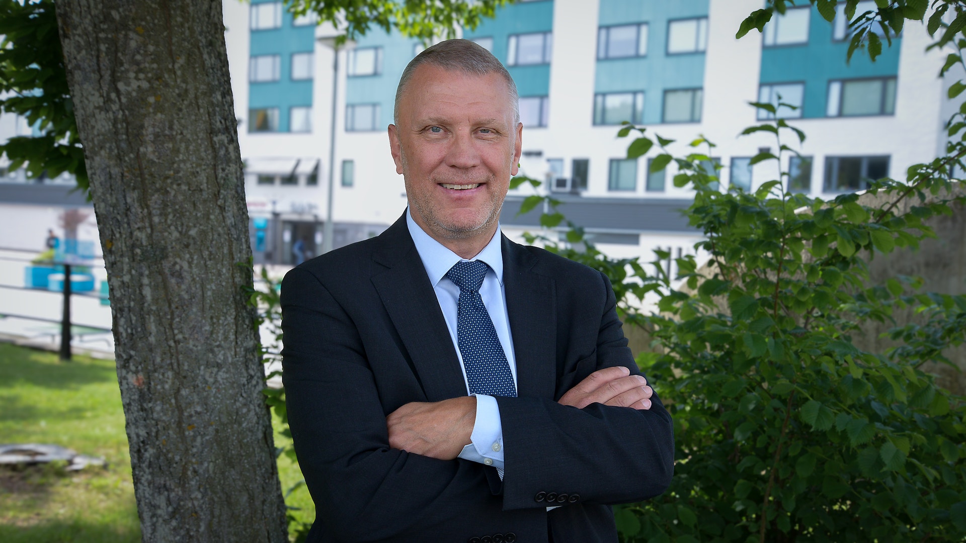 Porträtt av en man i mörk kostym och blå slips som står utomhus med armarna i kors. Modern bostadsbyggnad syns i bakgrunden.