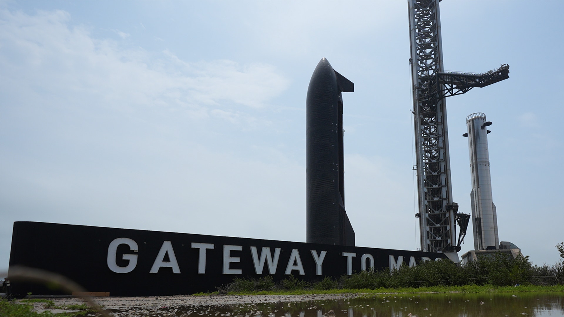 En stor svart metallskulptur av en raket med texten "Gateway to Mars" framför. Industriella kranar och konstruktioner syns i bakgrunden mot en molnig himmel.