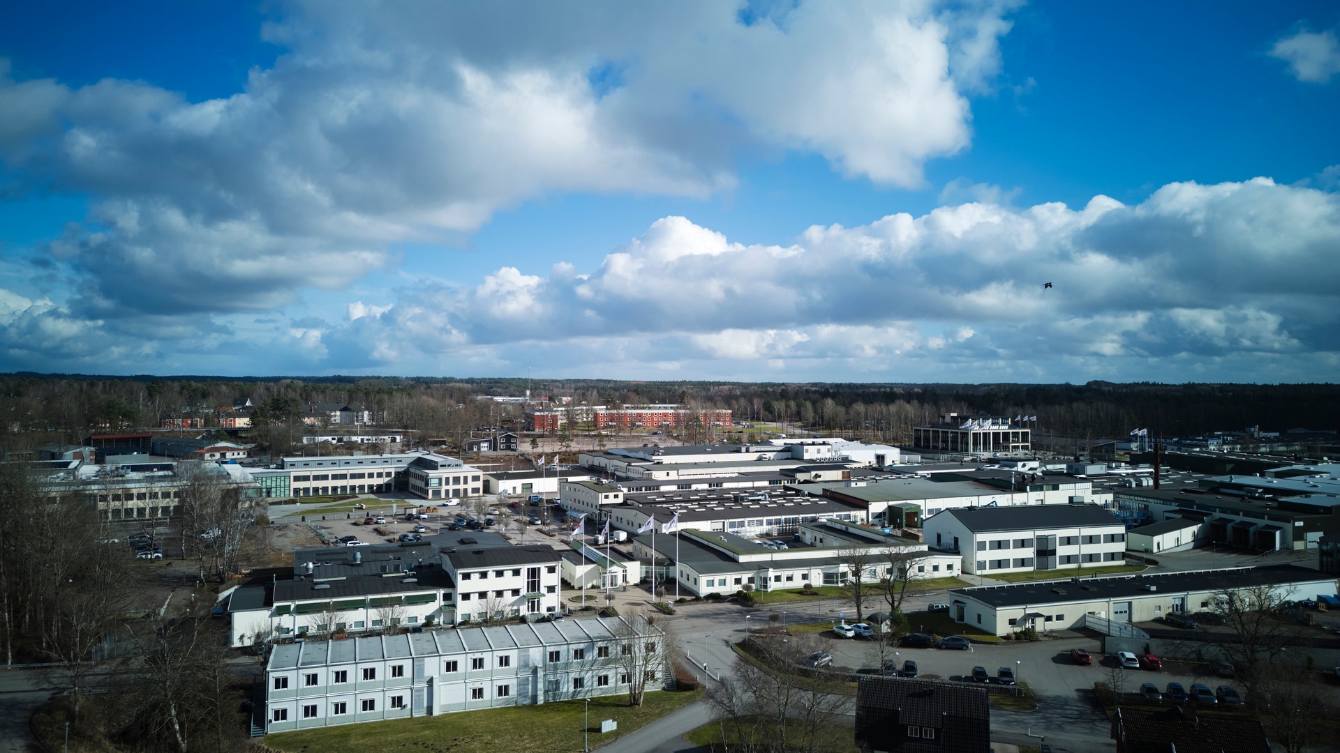 En äldre man med glasögon och blå kostym bär en orange säkerhetväst på en industriell arbetsplats med transportband och lagerhyllor i bakgrunden.