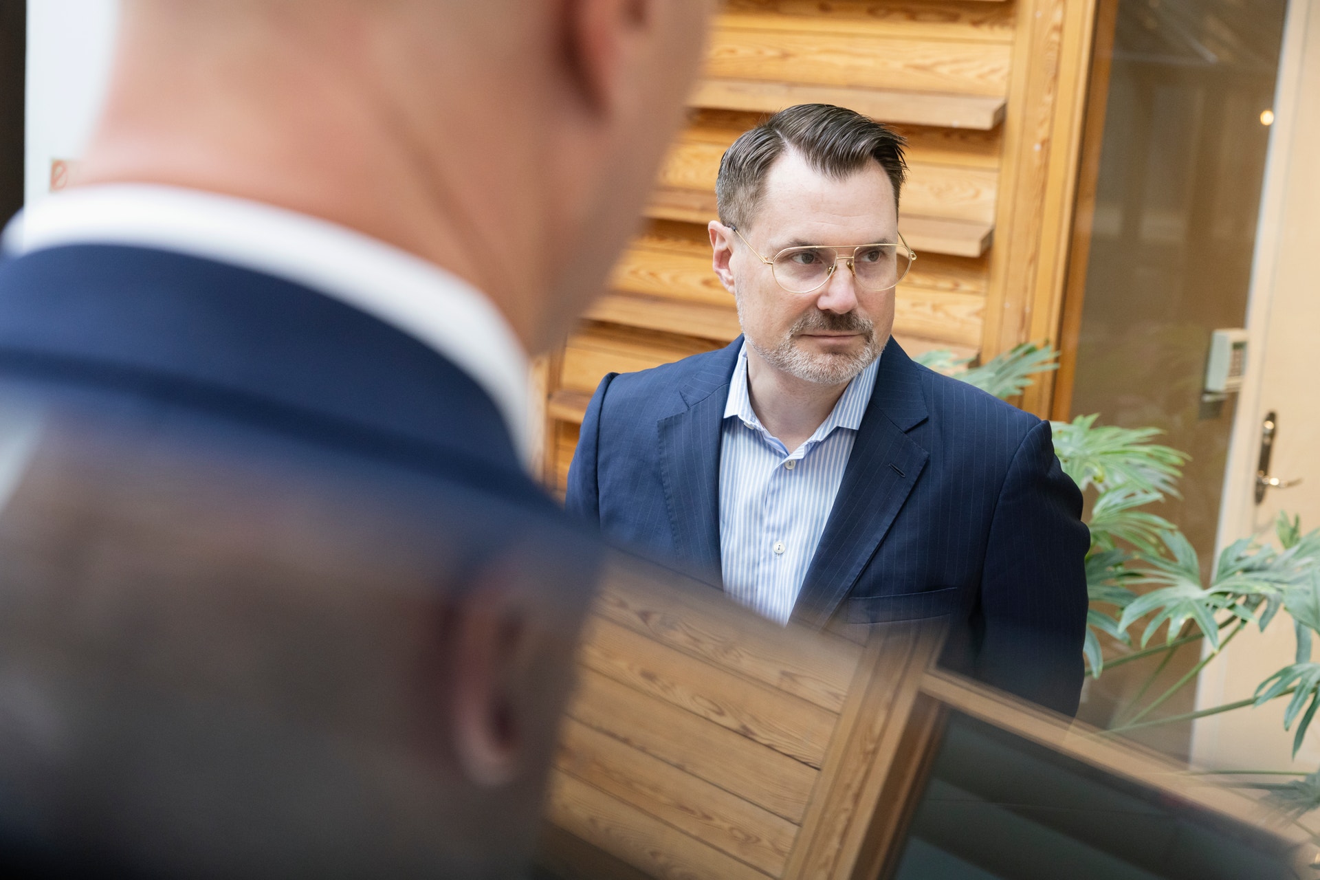 Två män i samtal inomhus. En man i mörkblå kavaj och ljusblå skjorta står i bakgrunden framför en vägg av trä.