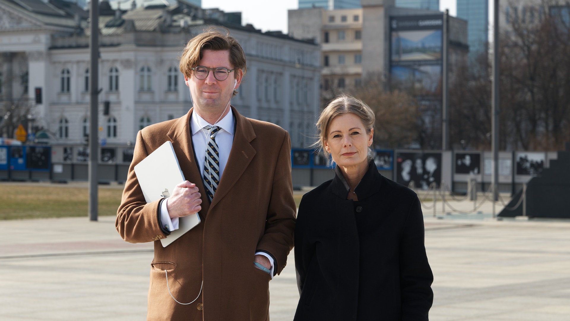 Två personer går tillsammans på en öppen plats framför klassiska byggnader