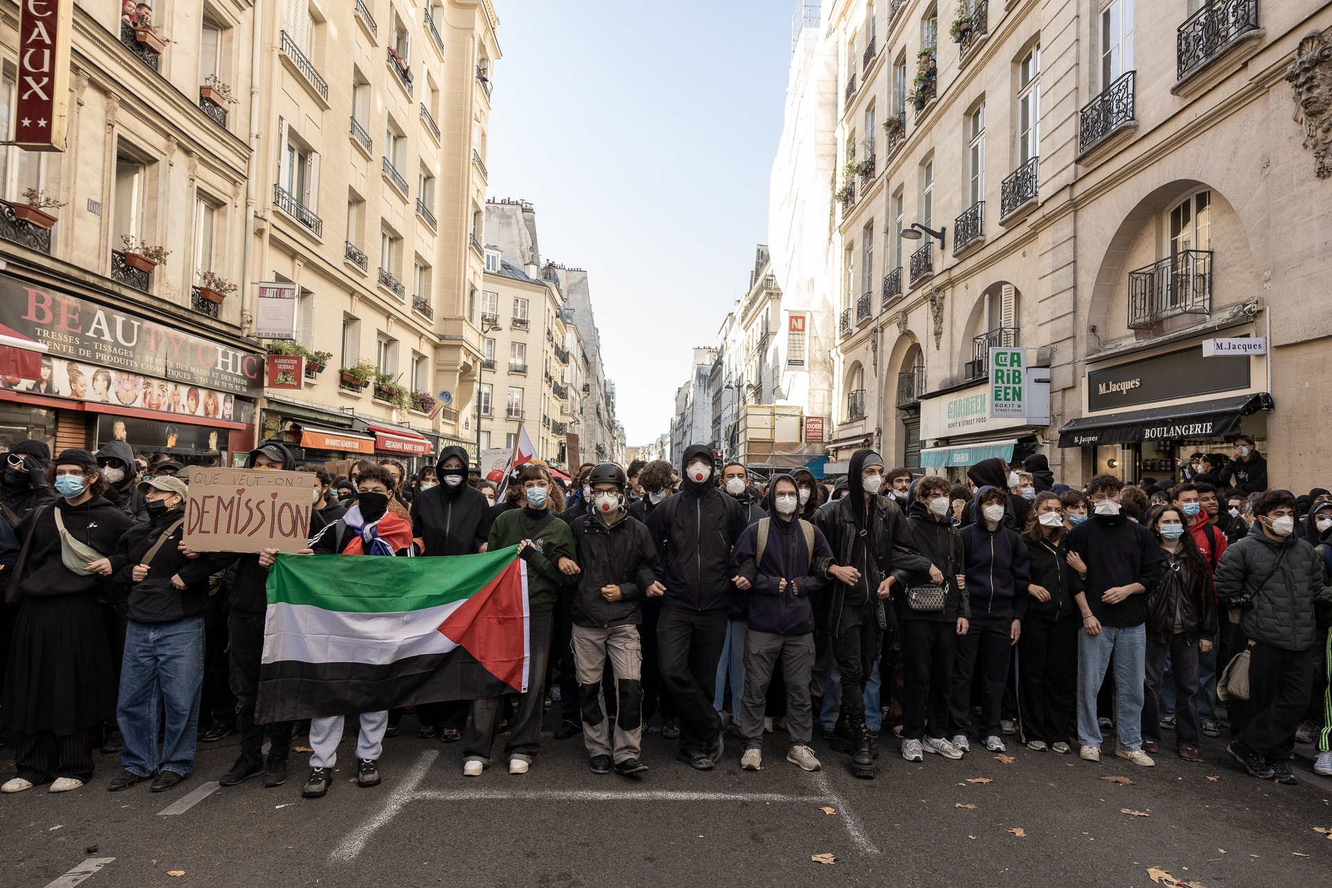 En stor folkmassa, många med munskydd och huvor, går tillsammans längs en gata i en stad. Längst fram bär några en palestinsk flagga och en skylt.