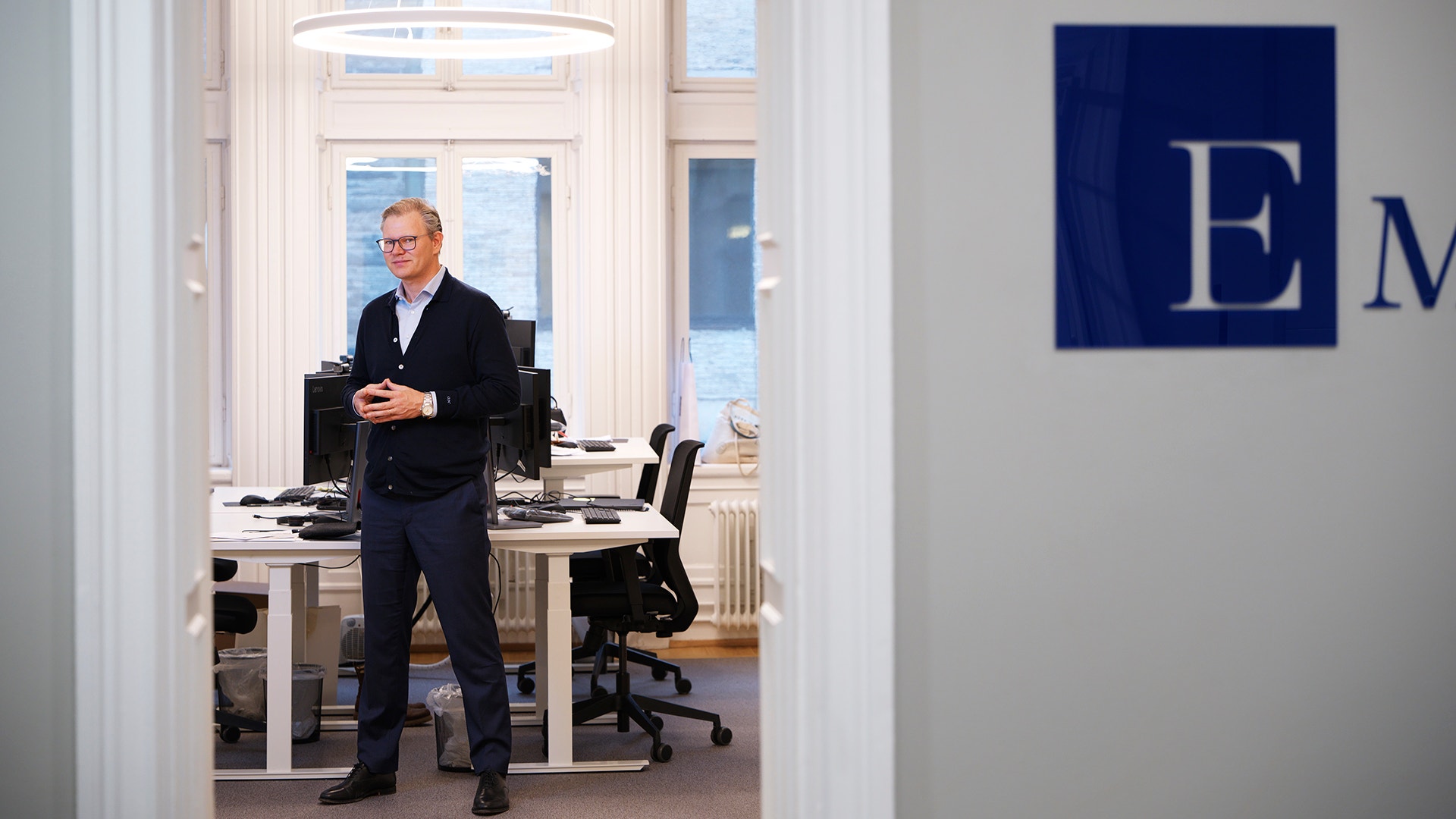 En man i mörk kofta och byxor står och ler i ett kontor, sett genom en dörröppning.