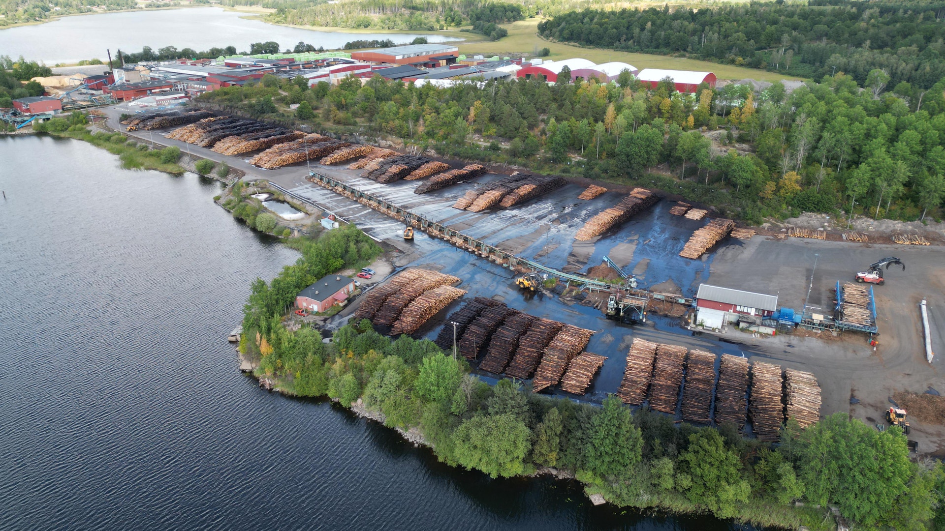 Flygfoto av ett sågverk vid en vattendrag med stora högar av timmer och industribyggnader omgivna av grönska.