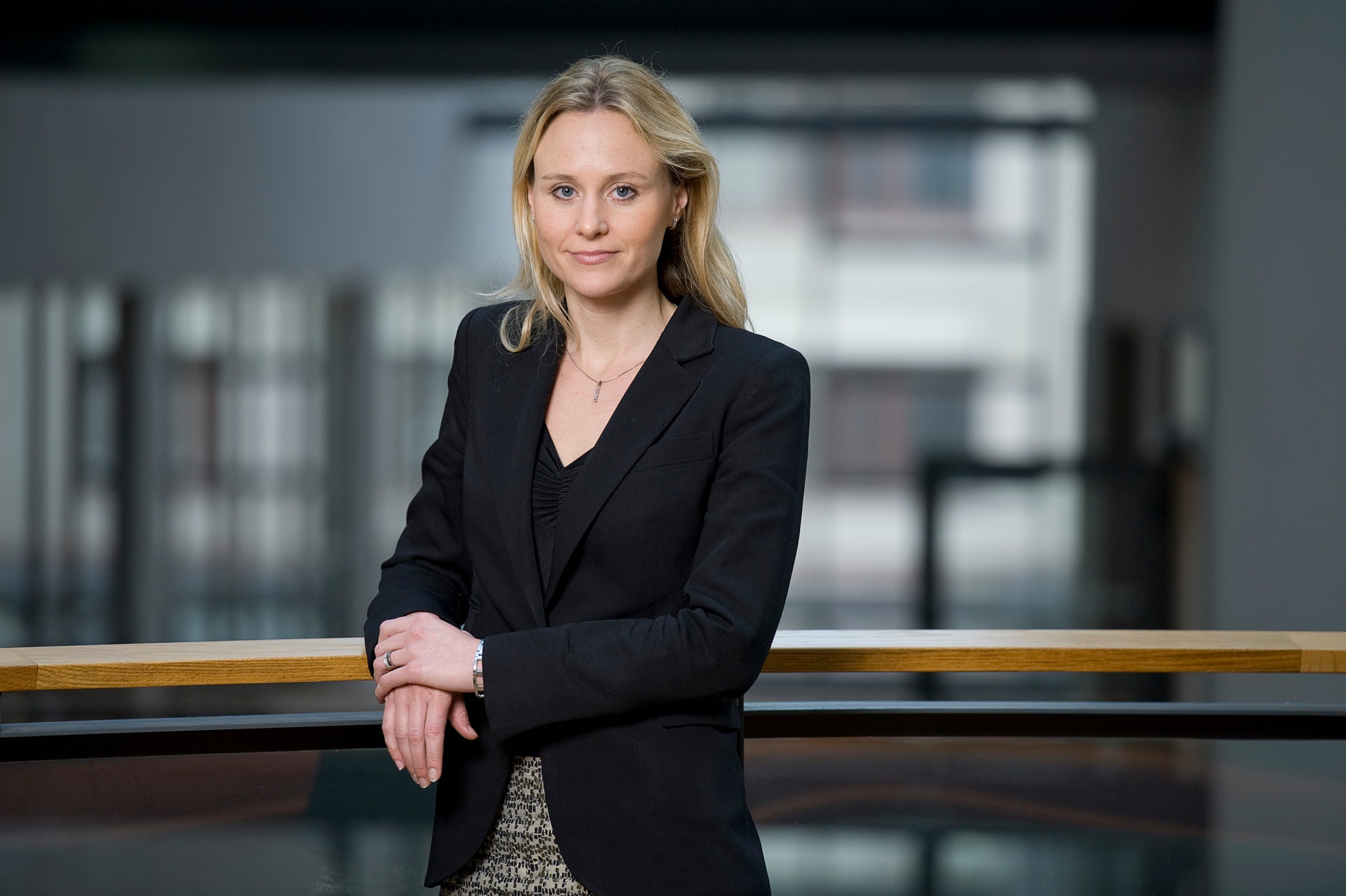 Porträtt av en kvinna i svart blazer som står vid ett räcke inomhus. Hon har blont hår och lutar sin arm mot räcket. Bakgrunden visar ett modernt kontorslandskap.