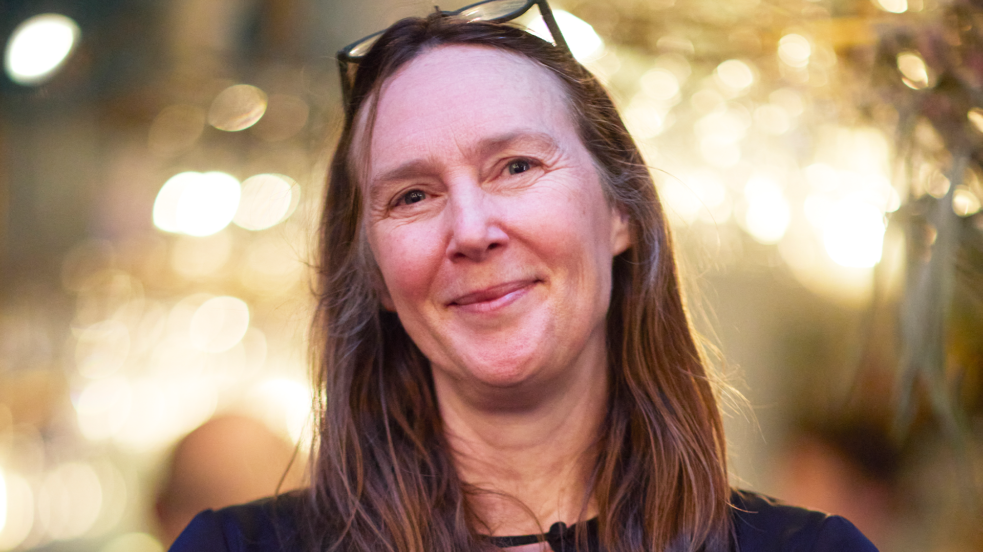 Porträttfoto av en leende kvinna med långt brunt hår mot en mjukt belyst bakgrund.