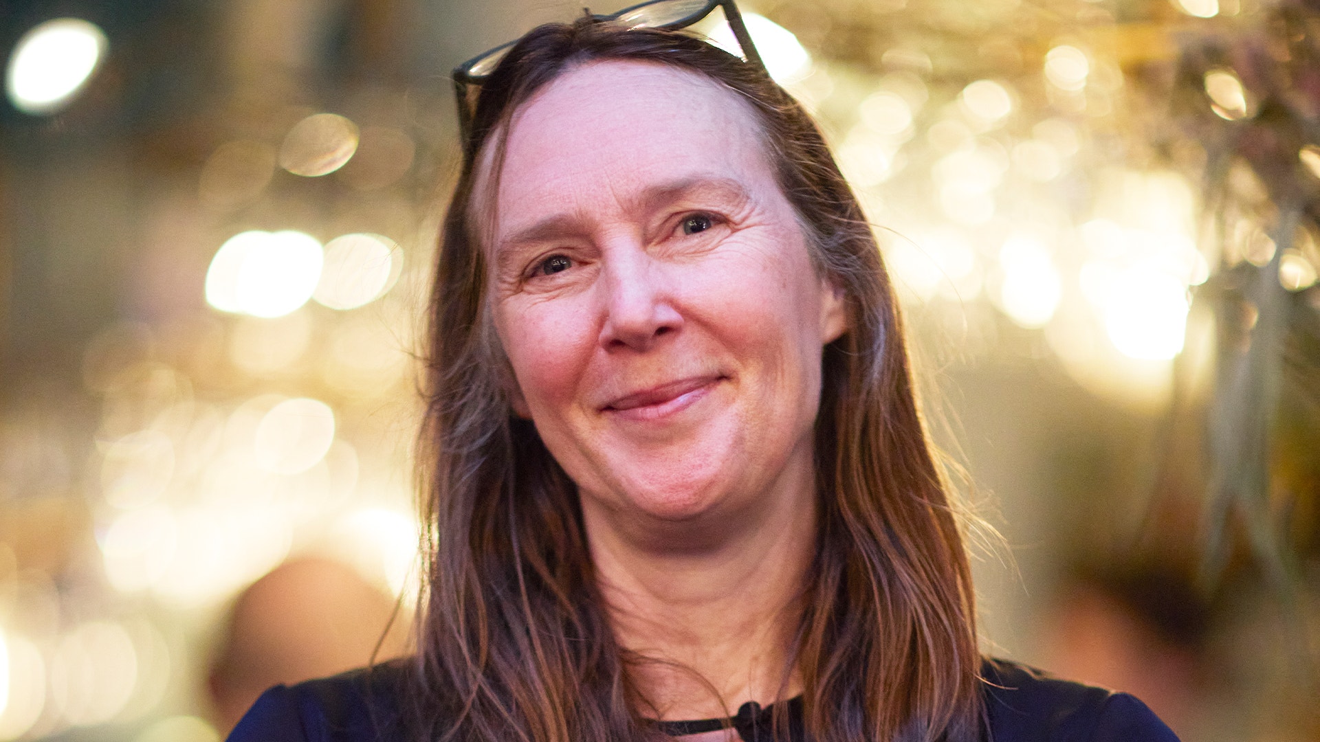 Porträttfoto av en leende kvinna med långt brunt hår mot en mjukt belyst bakgrund.