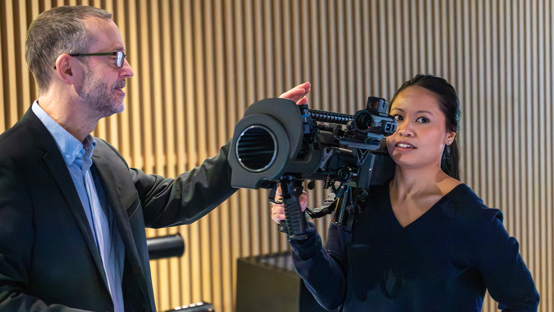 En man och en kvinna håller en professionell videokamera tillsammans i en studio med gul väggbakgrund.