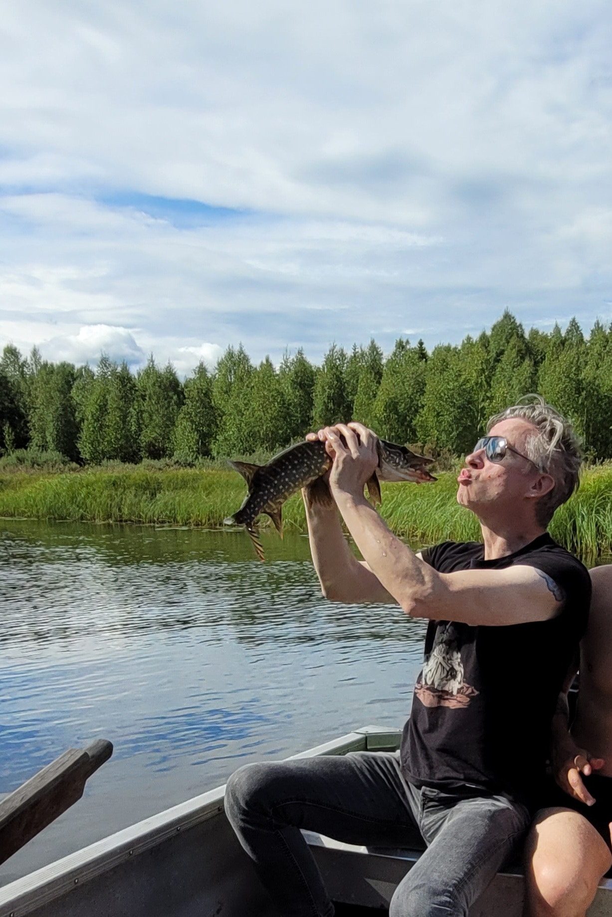 Man i båt som håller upp en fisk han har fångat. I bakgrunden syns en sjö, gröna ängar och barrskog under molnig himmel.