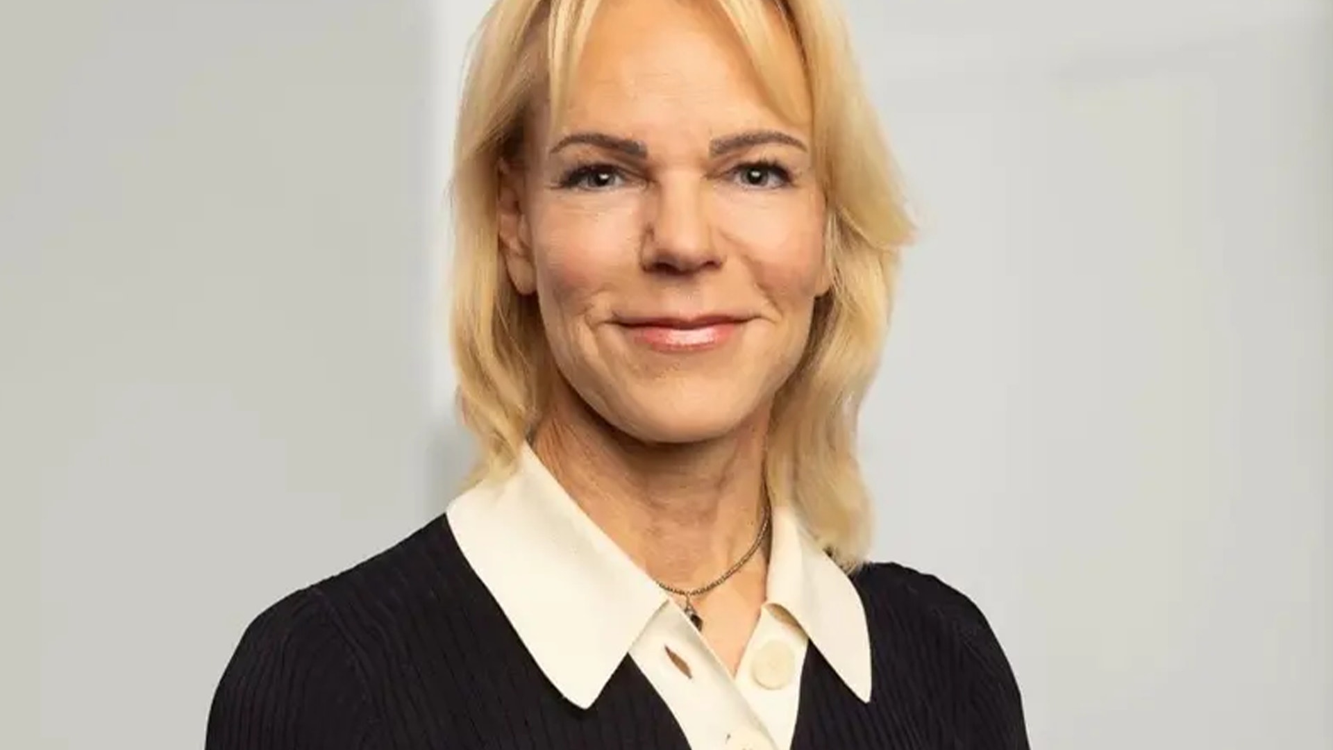 Porträtt av en kvinna med blont hår, svart jacka och vit krage som ler mot kameran.