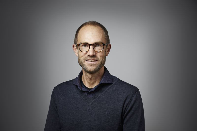 Porträtt av en leende man med glasögon, skäggstubb och mörk tröja framför grå bakgrund.