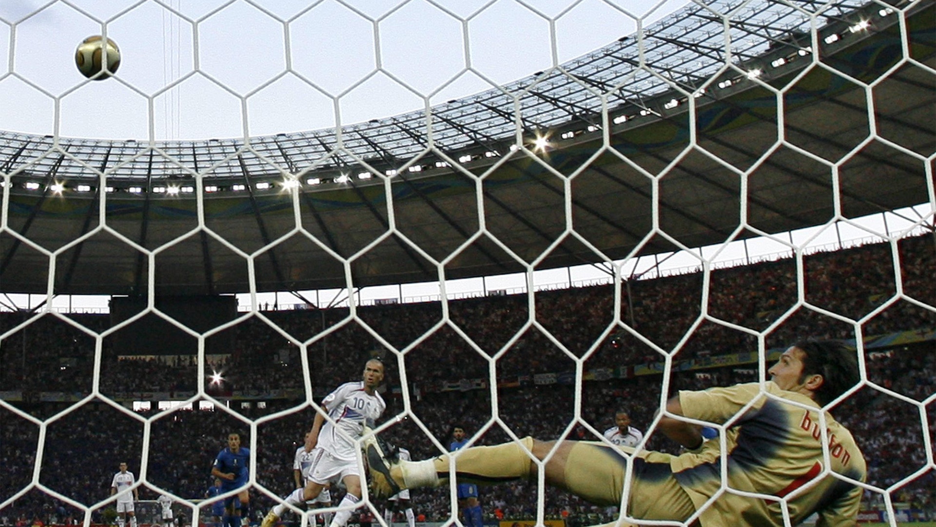 En fotbollsmatch där en boll är på väg in i målet. Bilden är tagen från bakom målnätet med målvakten i gult som försöker rädda på en stor stadion.