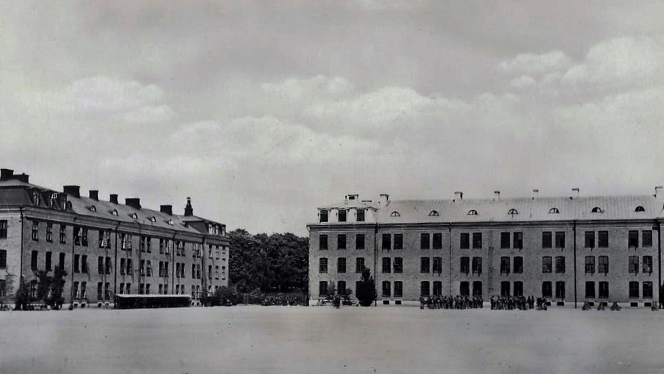 Svartvit historisk fotografi av två stora kasernbyggnader med klassisk arkitektur på en öppen plats.