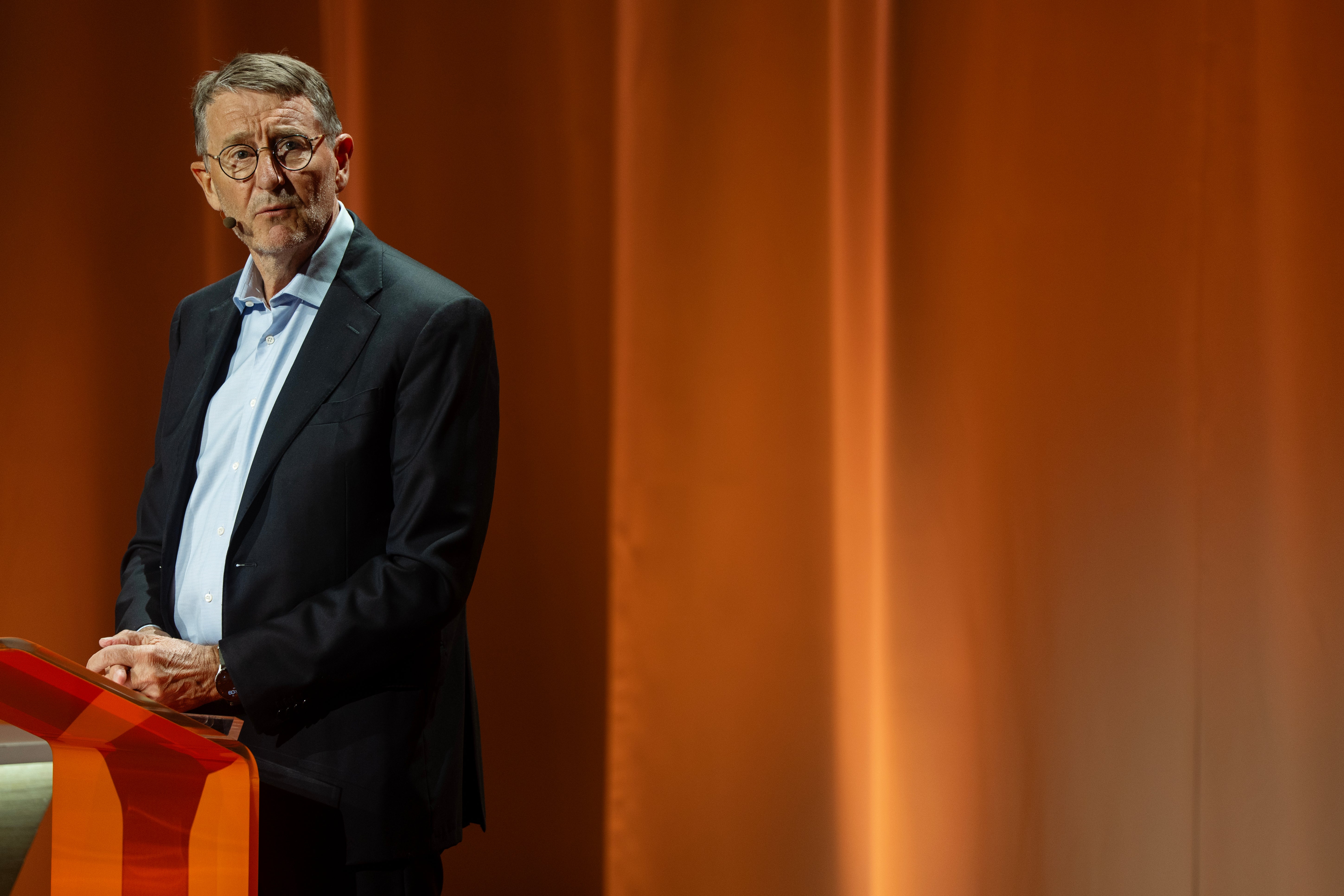 Man i glasögon och mörk kostym står vid ett rött podium på en scen med orange bakgrund.