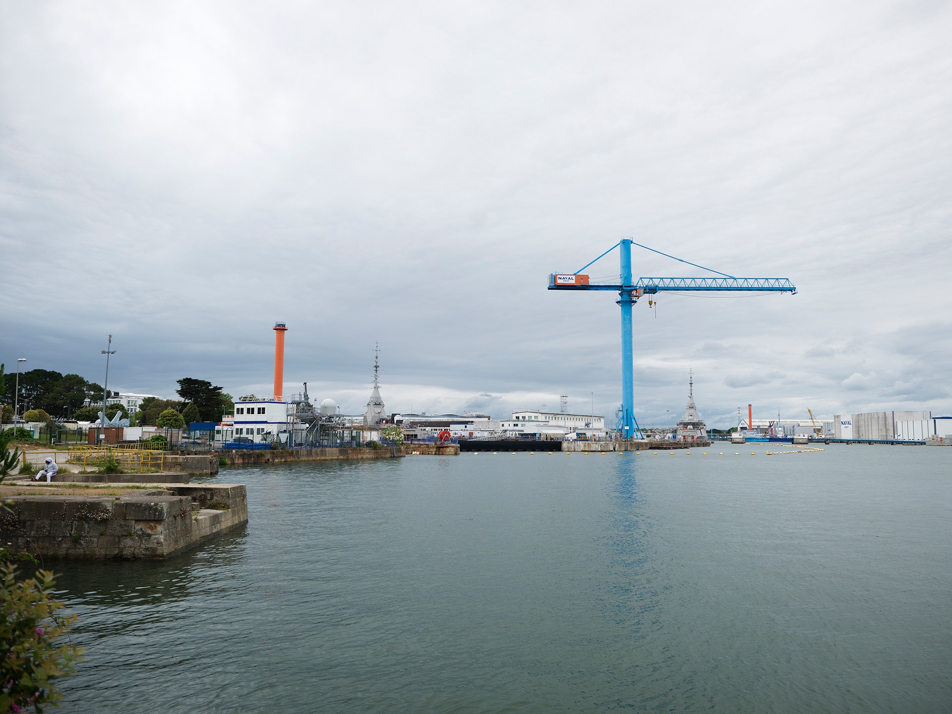 En stor blå lyftkran vid en industrihamn med vatten och industribyggnader i bakgrunden.