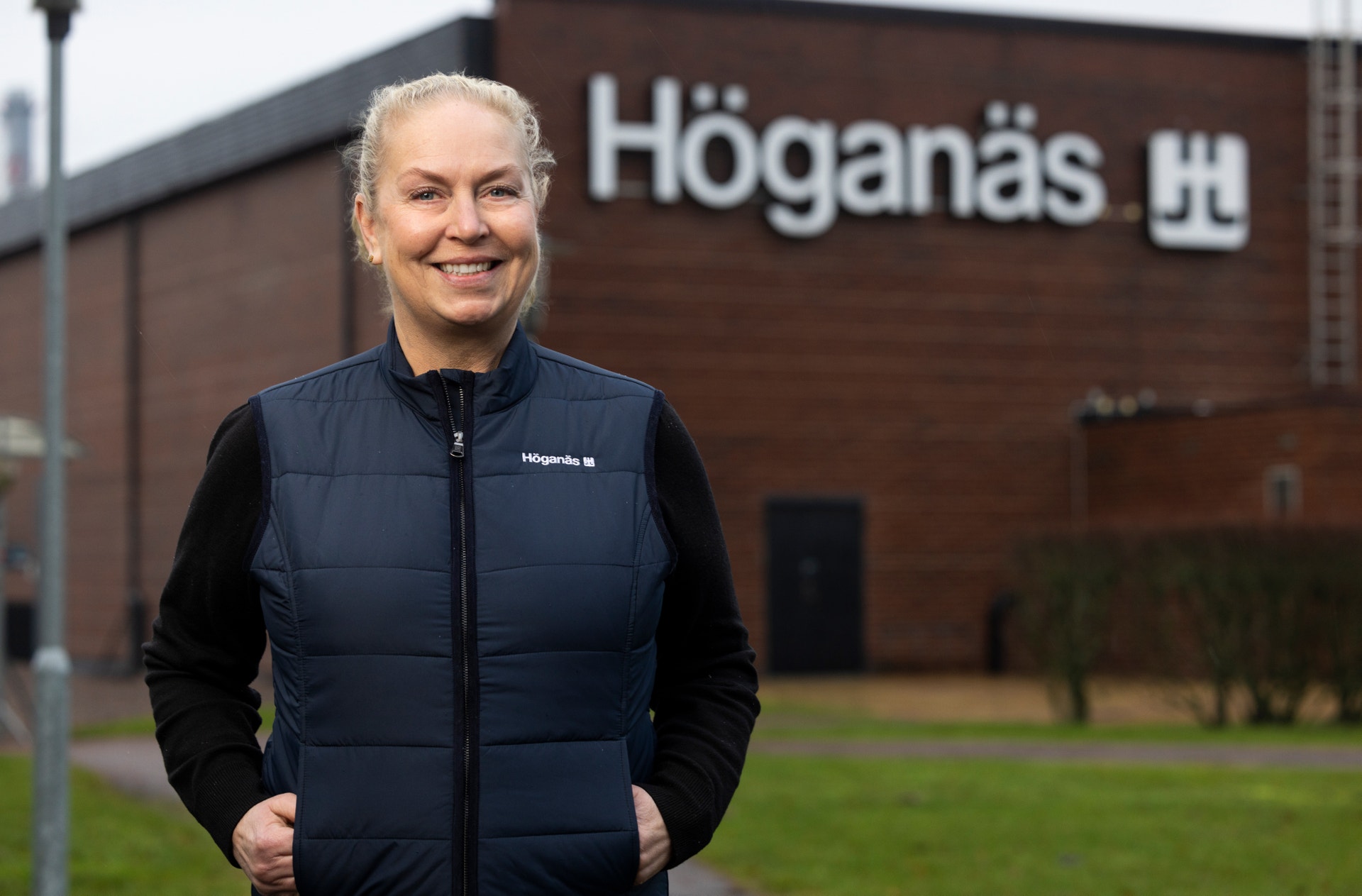 En leende kvinna står utomhus framför en fabriksbyggnad med texten Höganäs.