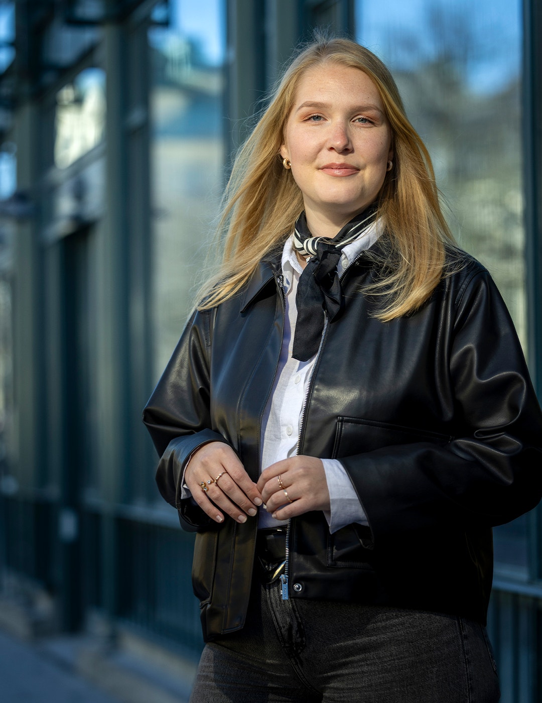 Porträtt av en ung kvinna med långt ljusbrunt hår, iklädd svart läderiacka och vit skjorta med svart halsduk, framför en urban bakgrund.