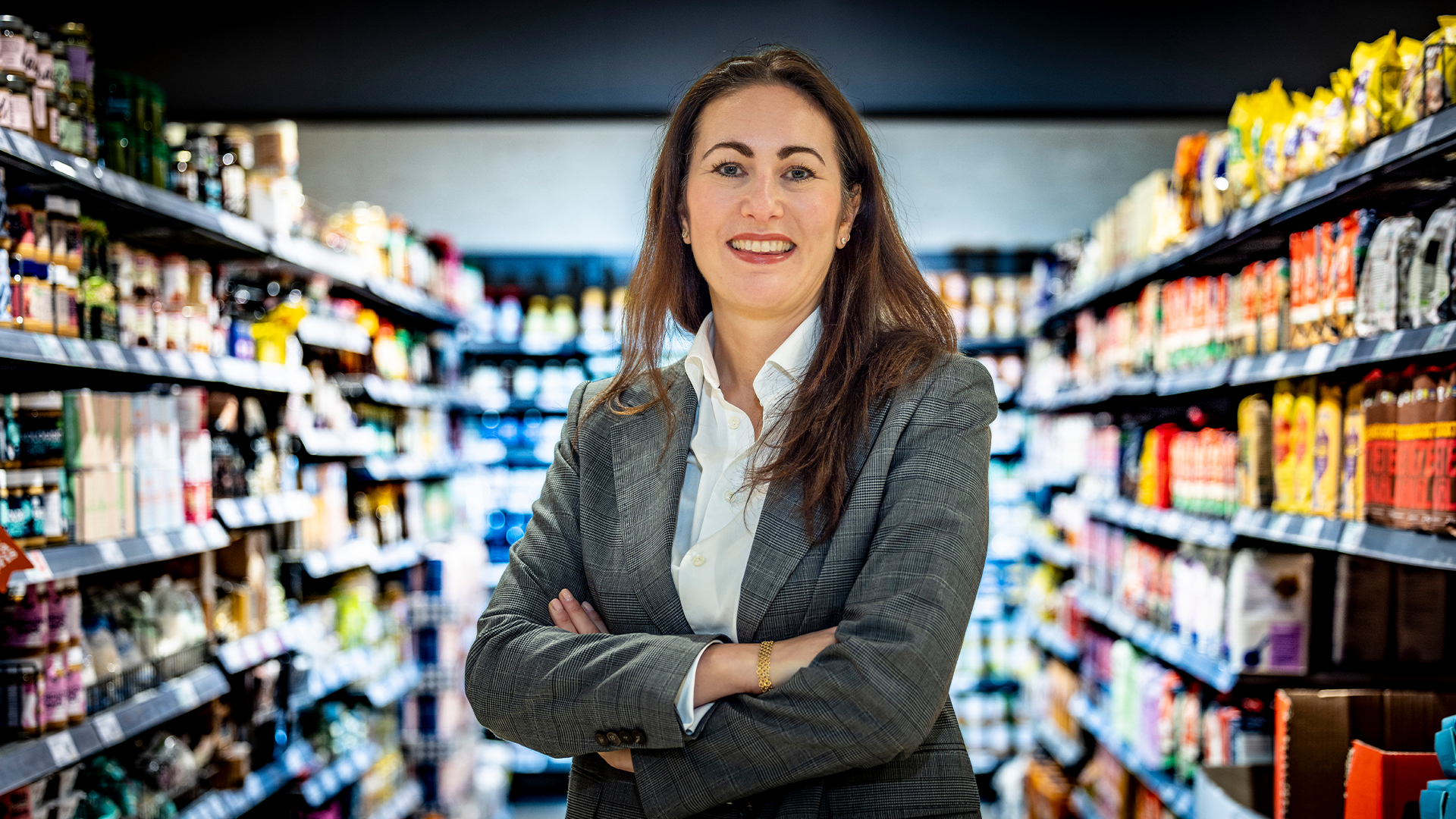 Porträtt av en leende kvinna i grå kostym och vit skjorta som står med armarna i kors framför hyllor fulla av matvaror i en stormarknad. Hon tittar direkt mot kameran med ett professionellt uttryck.