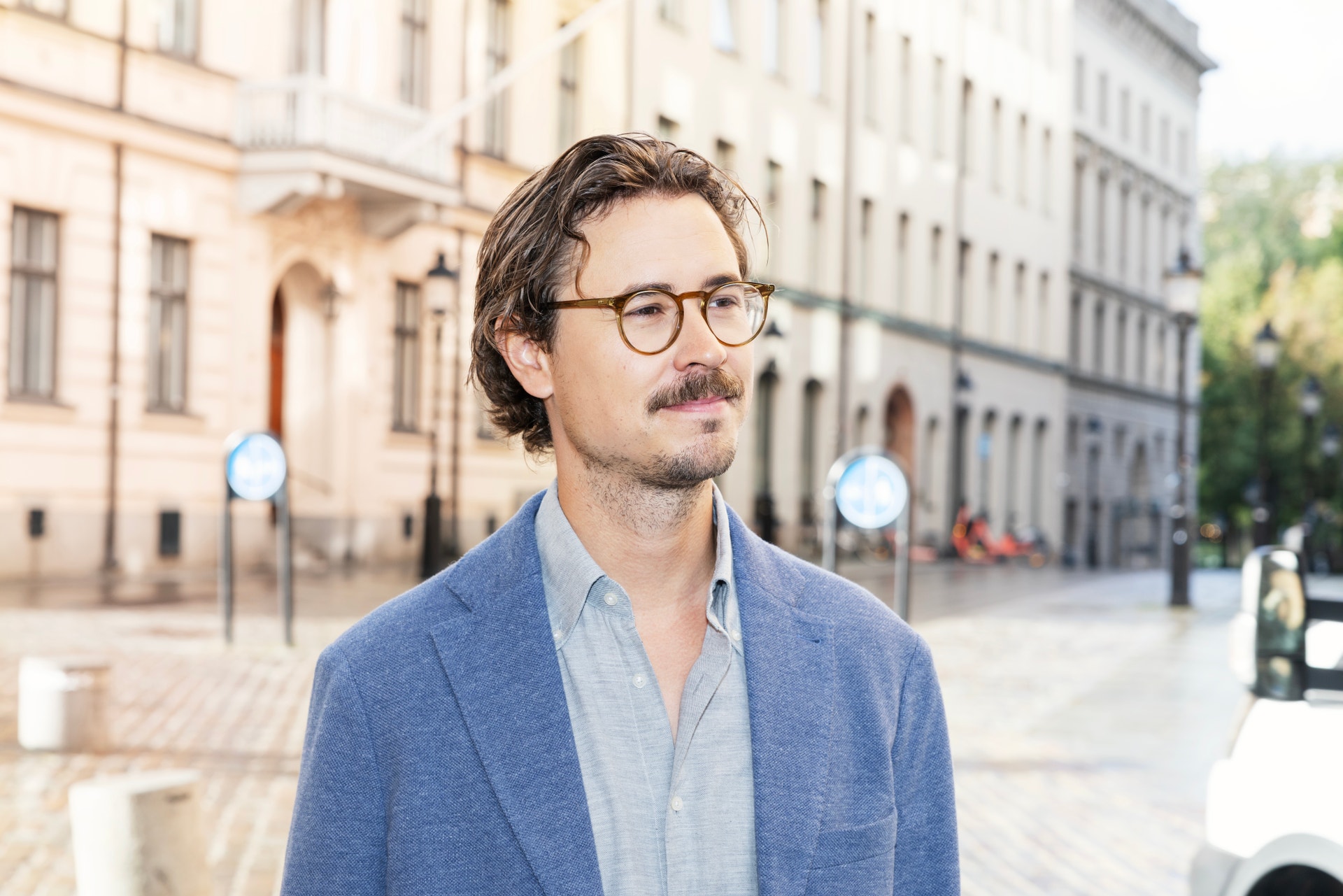 En man med glasögon och mustasch står på en gata i en stad, klädd i kavaj och skjorta.