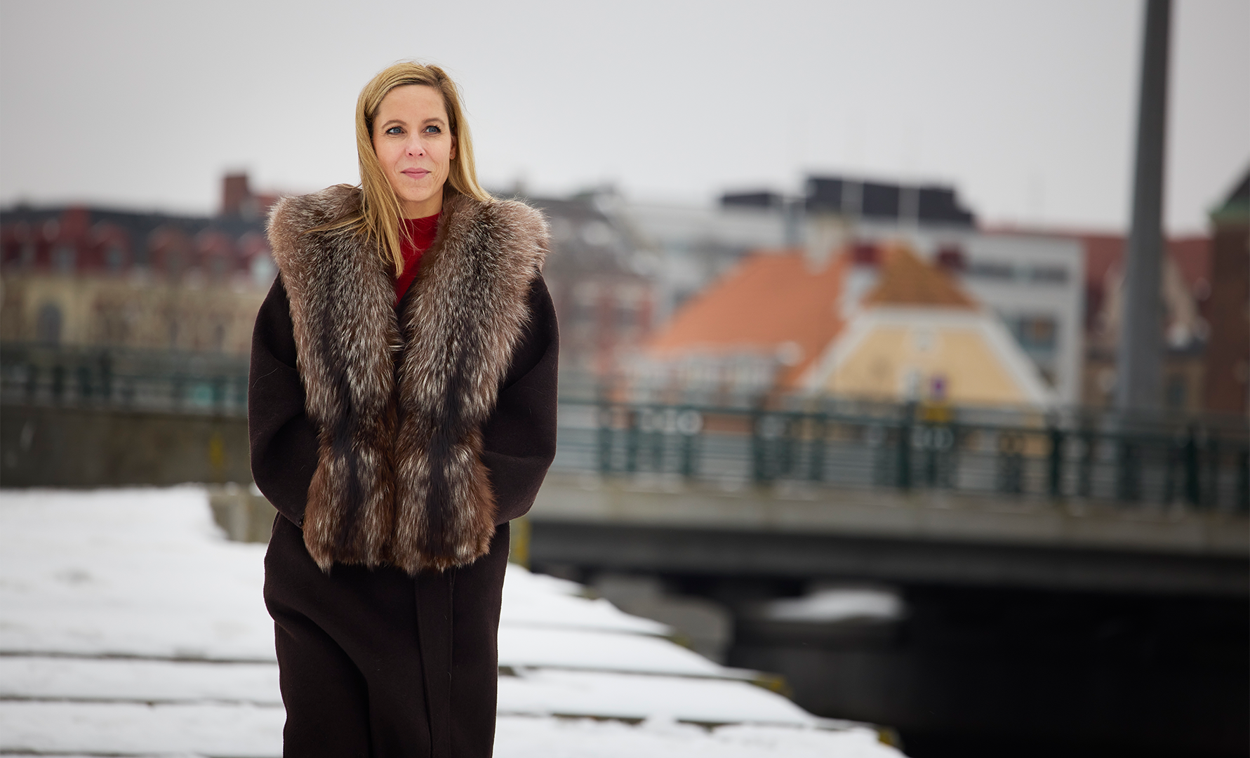 Porträtt av en kvinna i svart kappa med pälskrage, fotograferad vid en kanal i en stad.