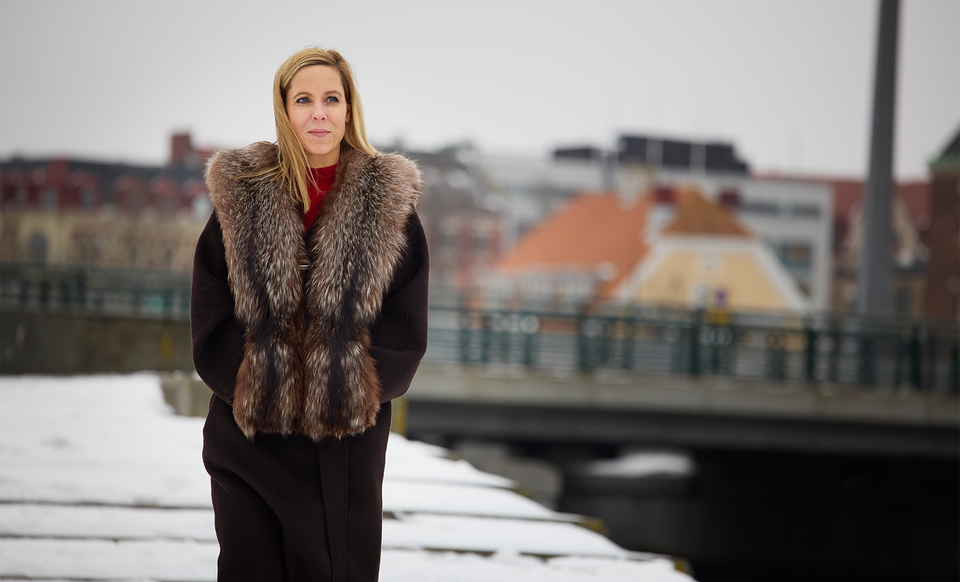 Porträtt av en kvinna i svart kappa med pälskrage, fotograferad vid en kanal i en stad.