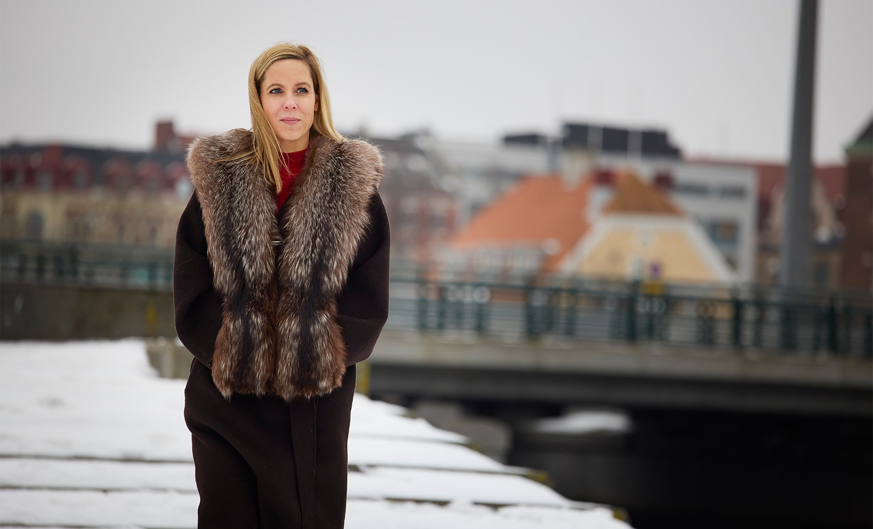 Porträtt av en kvinna i svart kappa med pälskrage, fotograferad vid en kanal i en stad.