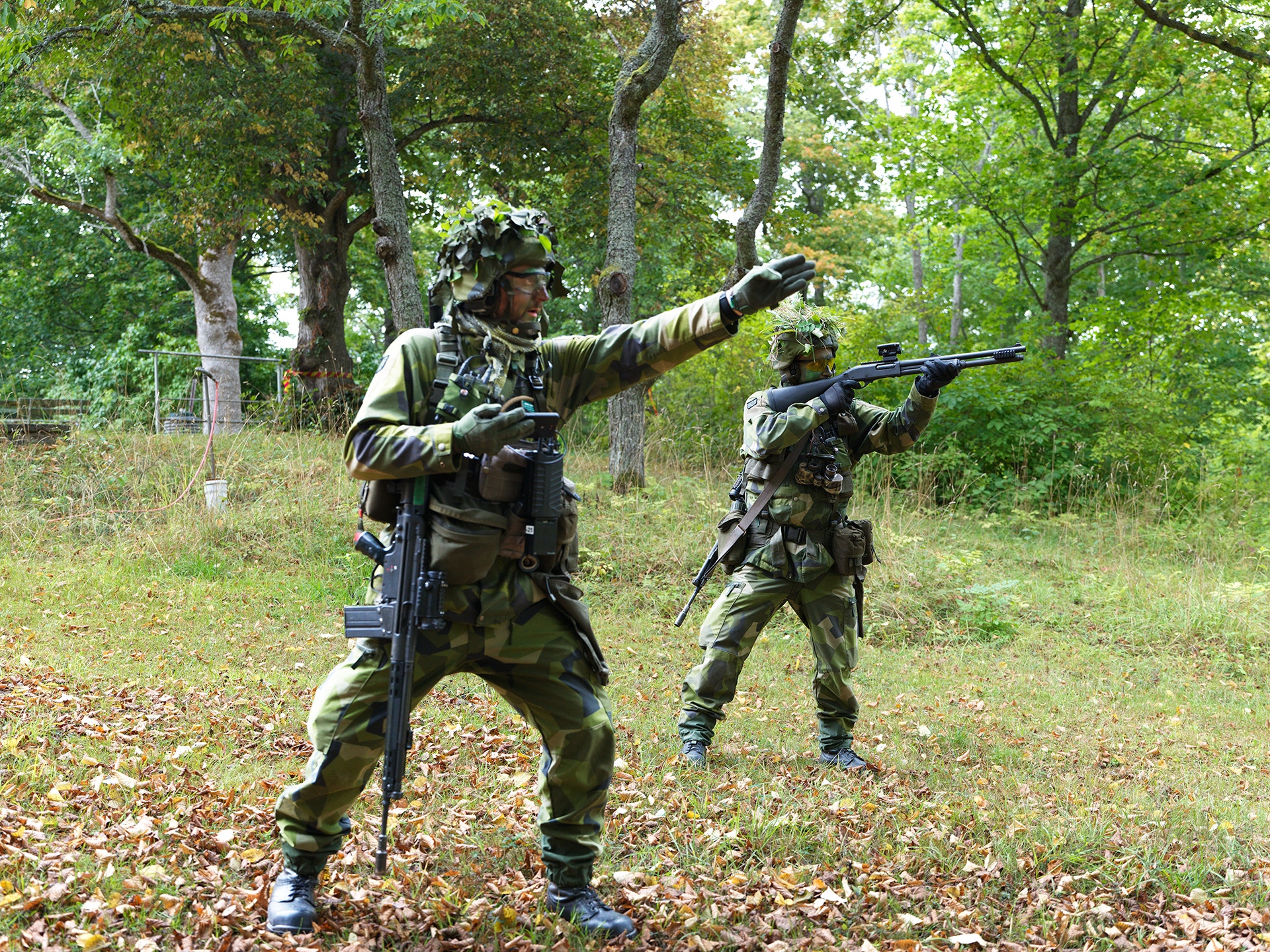 Två soldater i kamouflageuniform övar i skogsterräng, en pekar med handen och en siktar med sitt vapen.