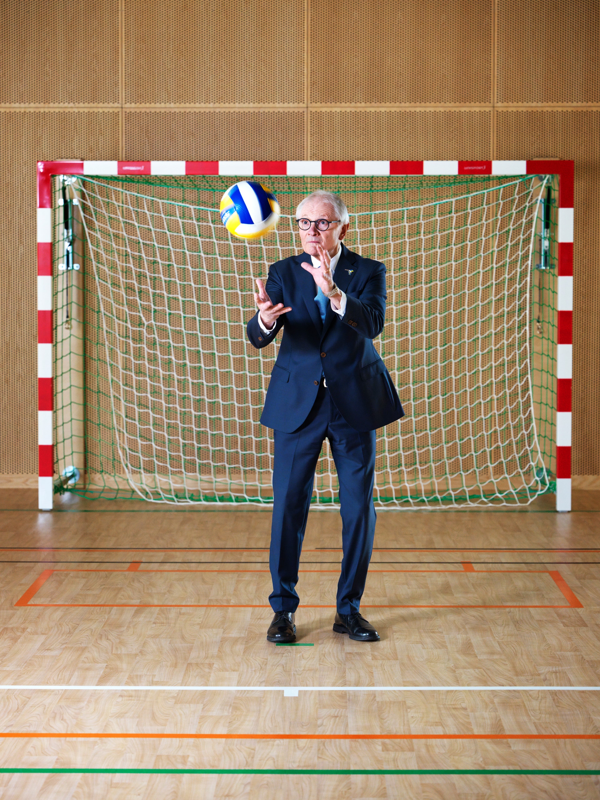 Man i mörkblå kostym kastar en handboll framför ett rött och vitt handbolls-mål inomhus.
