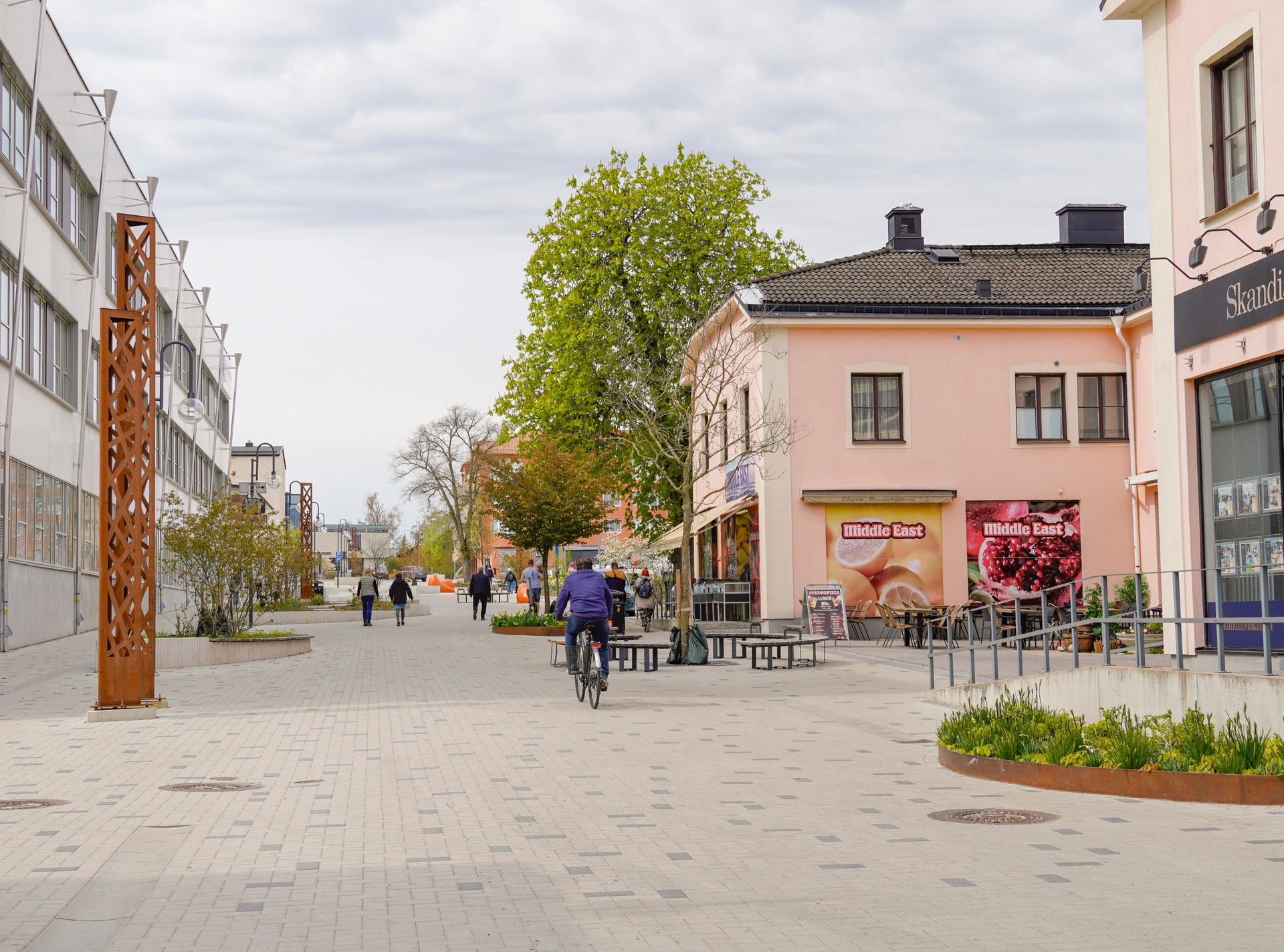 Finspångs centrum med ett öppet torg omgivet av klassiska byggnader, träd och butiker. Några personer promenerar på det stenlagda torget.