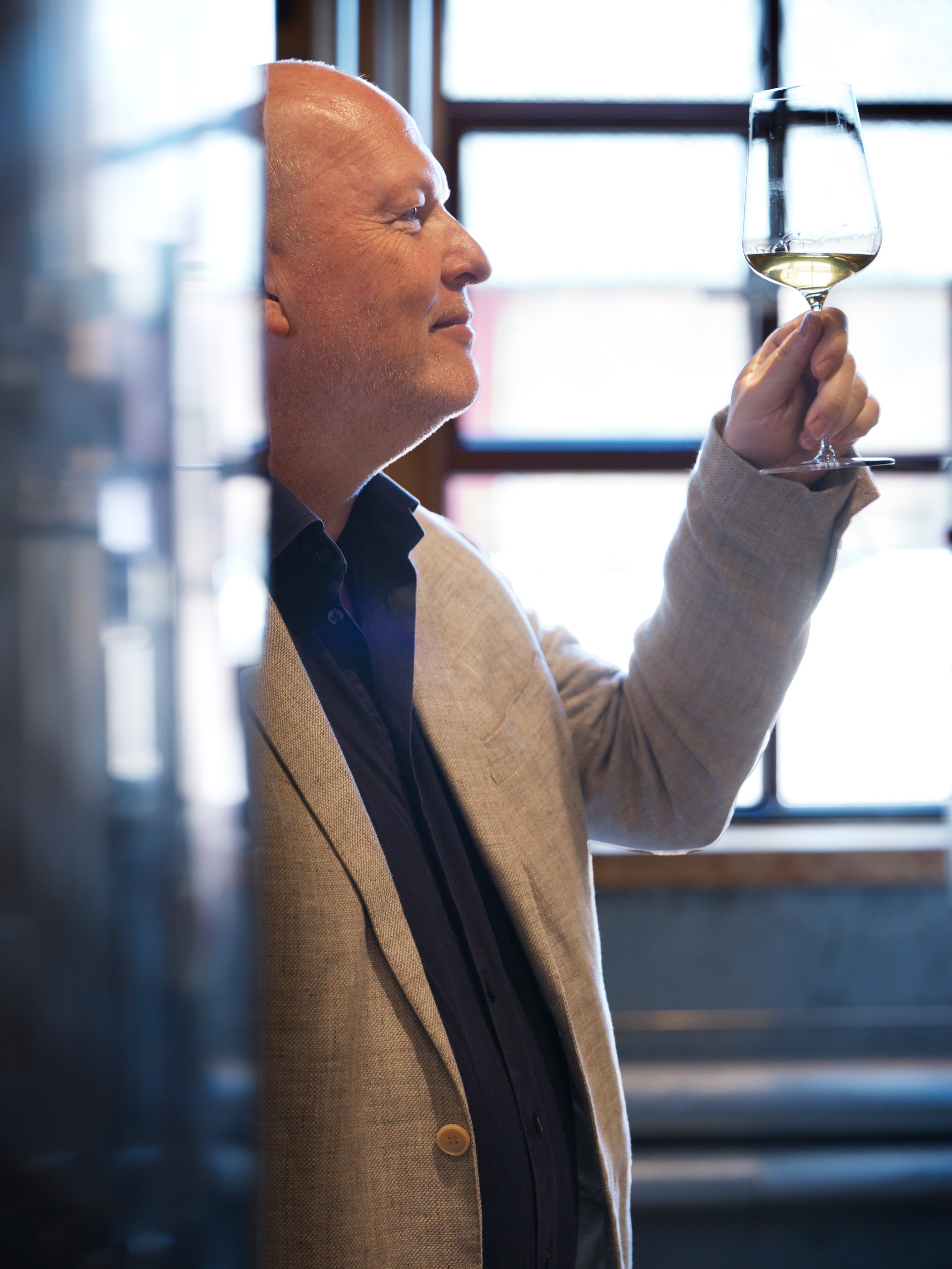 Man i ljus kavaj håller upp ett vinglas med vitt vin mot ljuset för att inspektera det. Han står vid ett fönster med stadsvy i bakgrunden.