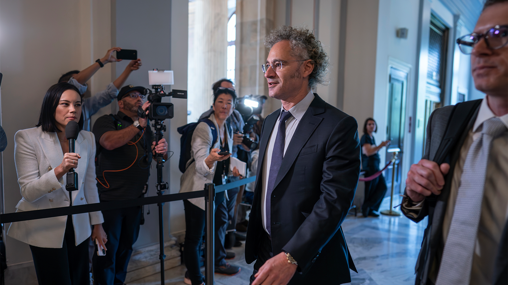 Alex Carp går genom entrén till Senaten i New York. Han omringas av reportrar.