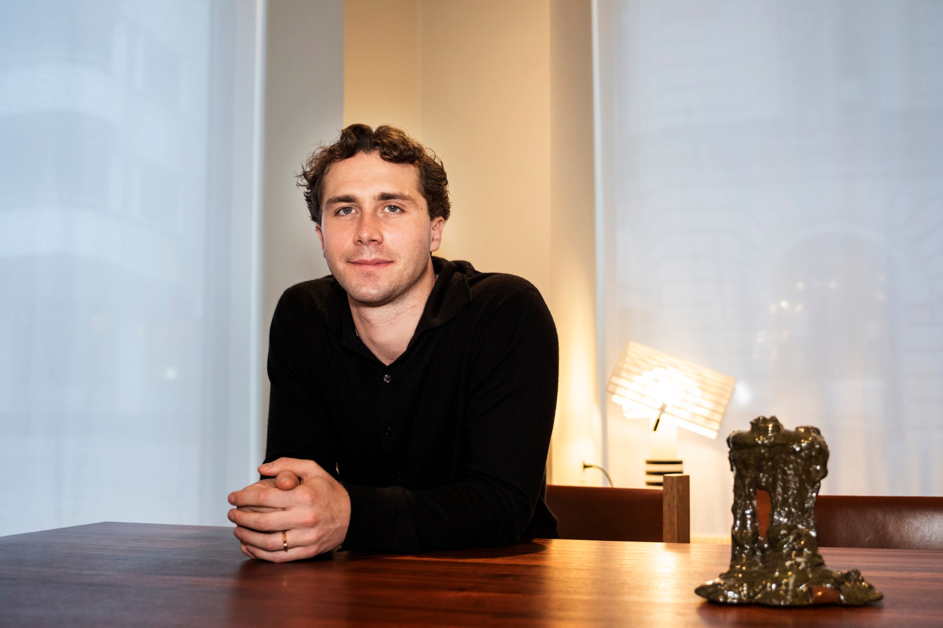 Man sitter vid ett bord och tittar mot kameran. Varmt ljus från en lampa bakom honom skapar en mysig atmosfär.