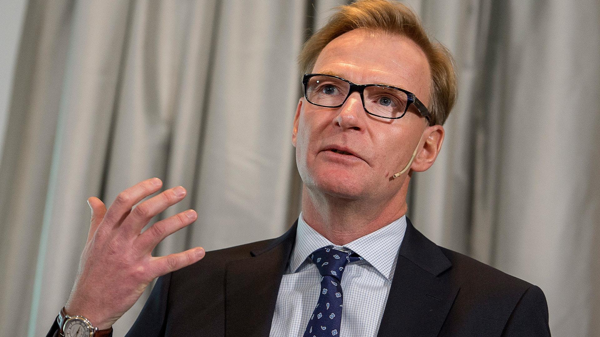 Man i affärskläder med glasögon gestikulerar medan han talar.