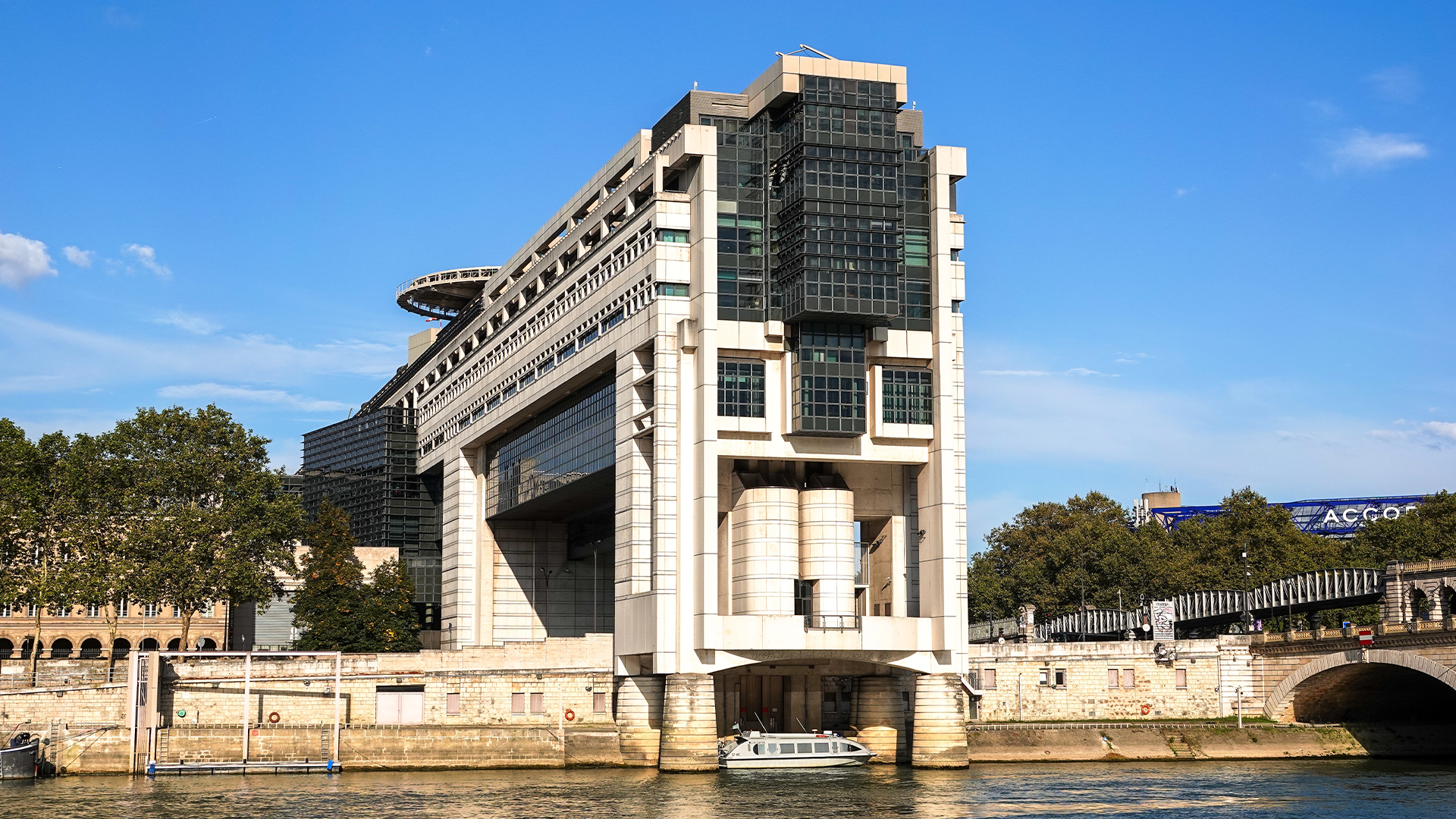 En stor modernistisk byggnad med vita och mörka fasader vid en flodkaj, med träd i bakgrunden och blå himmel.
