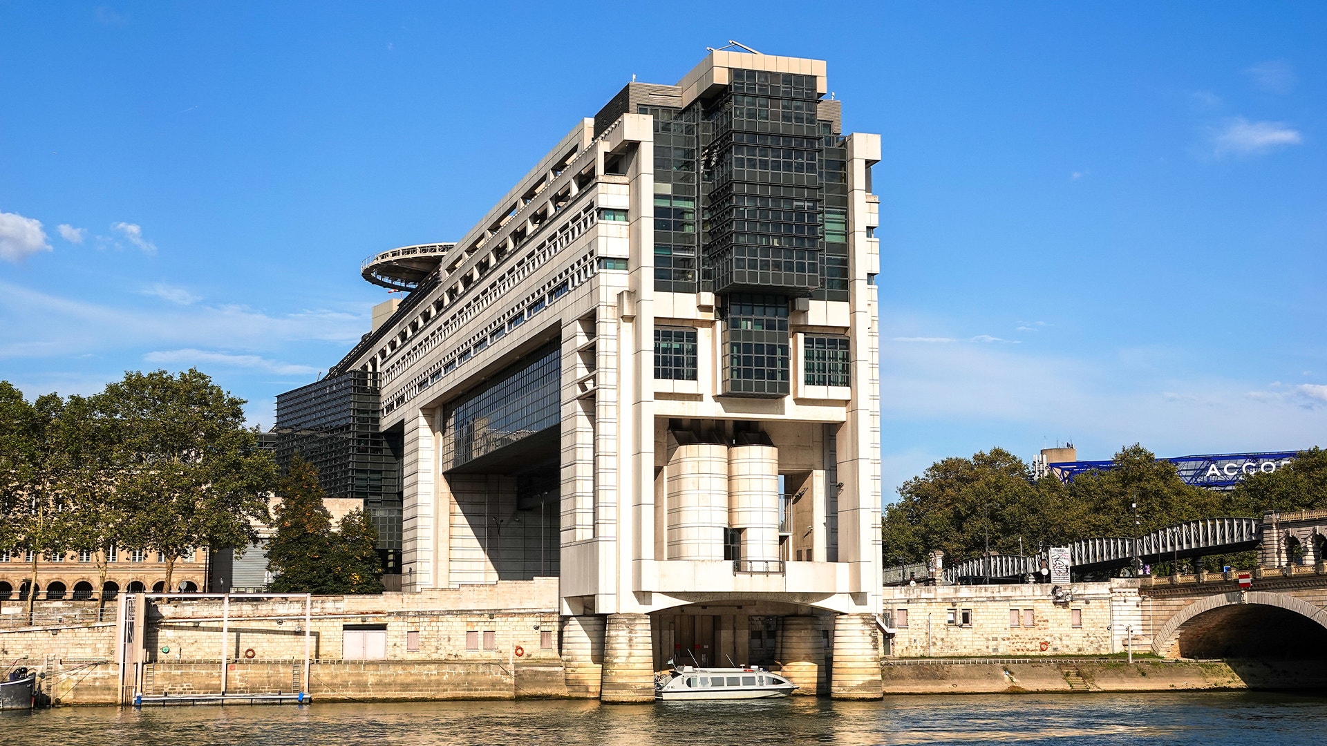 En stor modernistisk byggnad med vita och mörka fasader vid en flodkaj, med träd i bakgrunden och blå himmel.