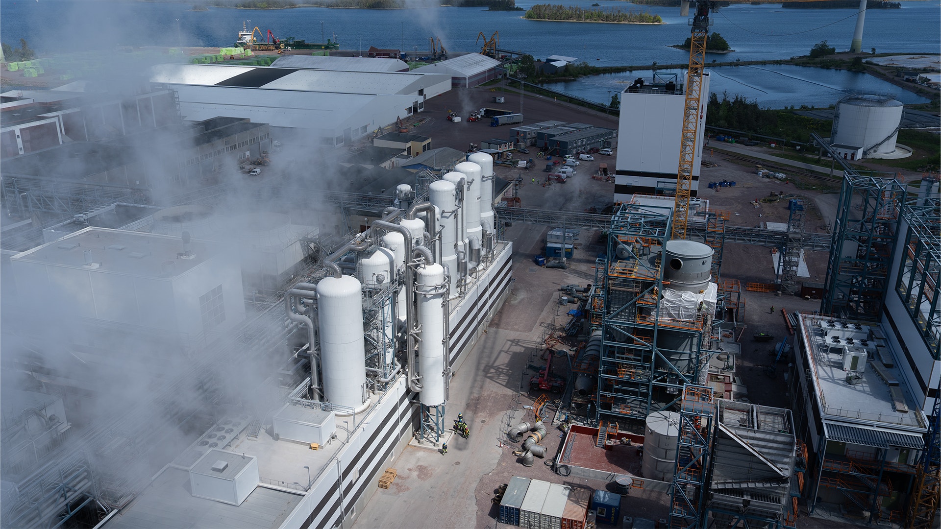 Luftfoto av en industrianläggning vid vatten med stora vita lagertankar, ånga som stiger upp, byggkran och fabriksstrukturer.