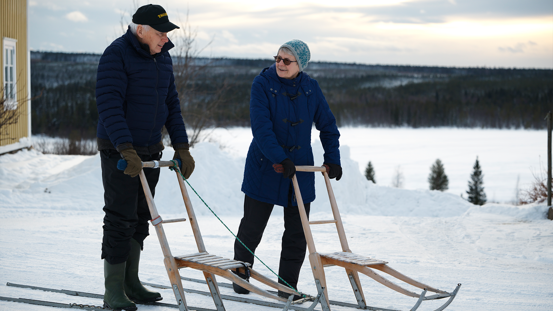 Två personer i vinterkläder står med skidor i snölandskapet med en frusen sjö och skog i bakgrunden.