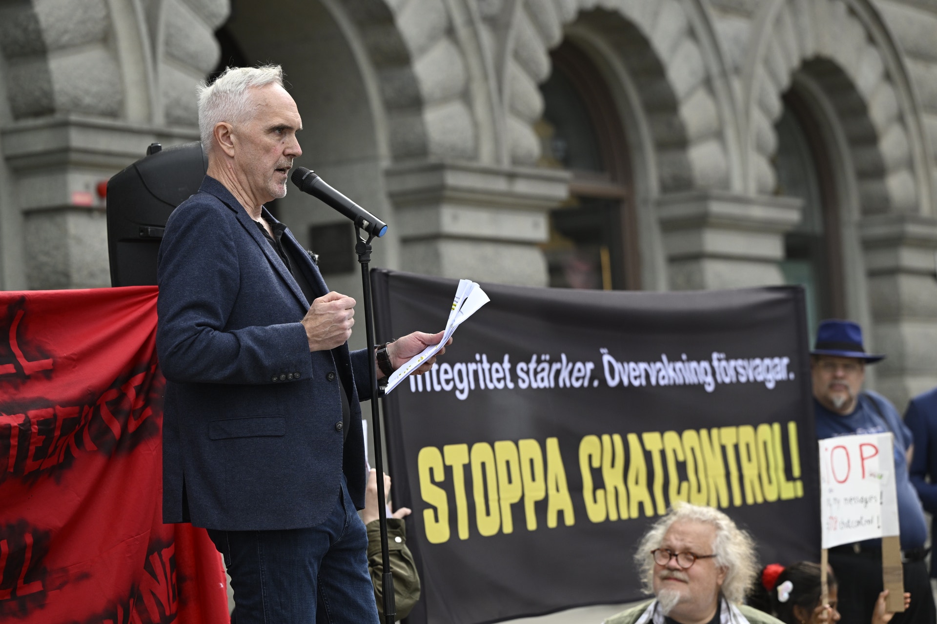 En man med vitt hår talar vid en mikrofon under en demonstration. I bakgrunden syns en banner med texten 'STOPPA CHATCONTROL' och en klassisk byggnad.