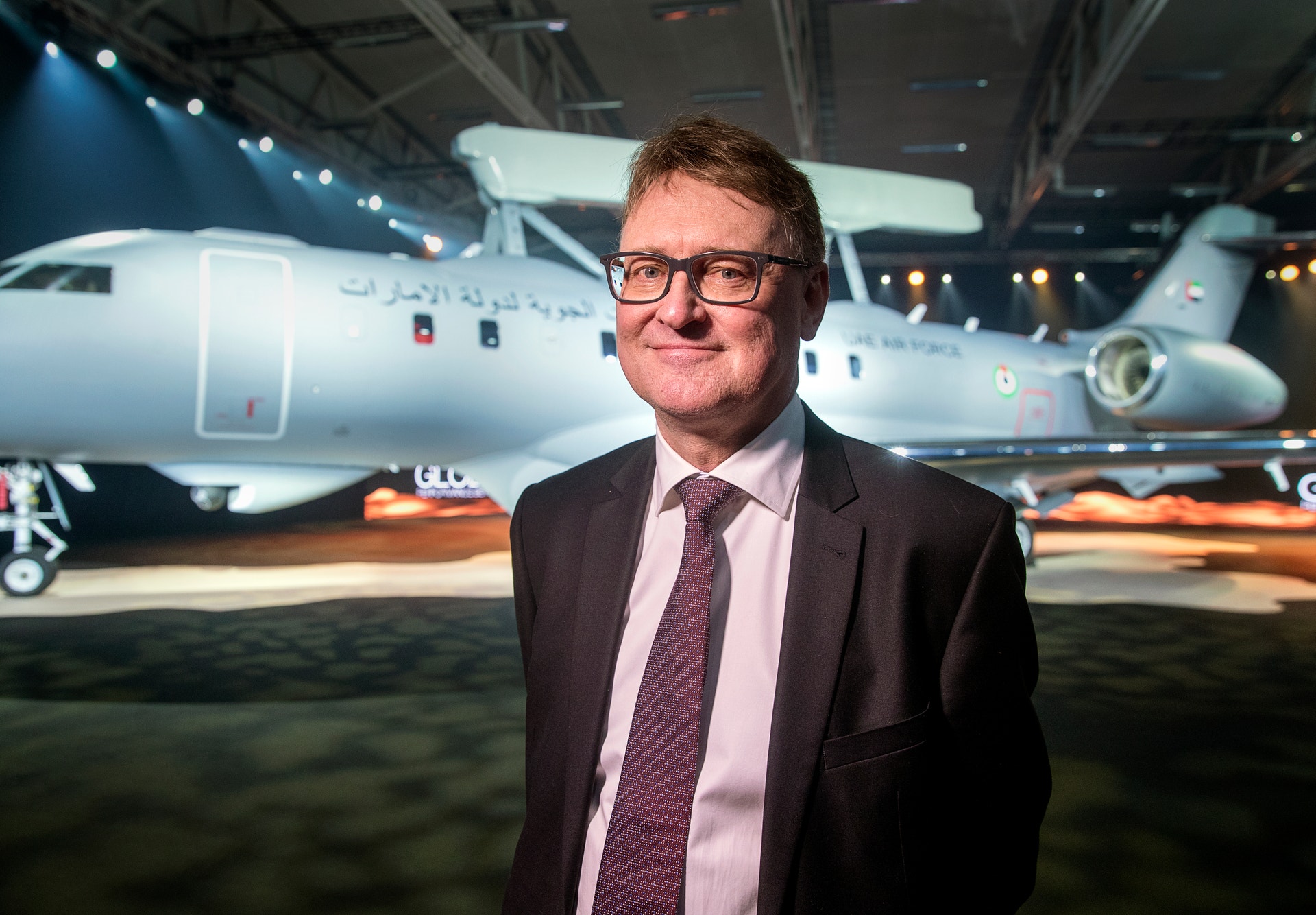 Leende man med glasögon, mörk kostym och slips står framför ett stort grått flygplan i en hall.