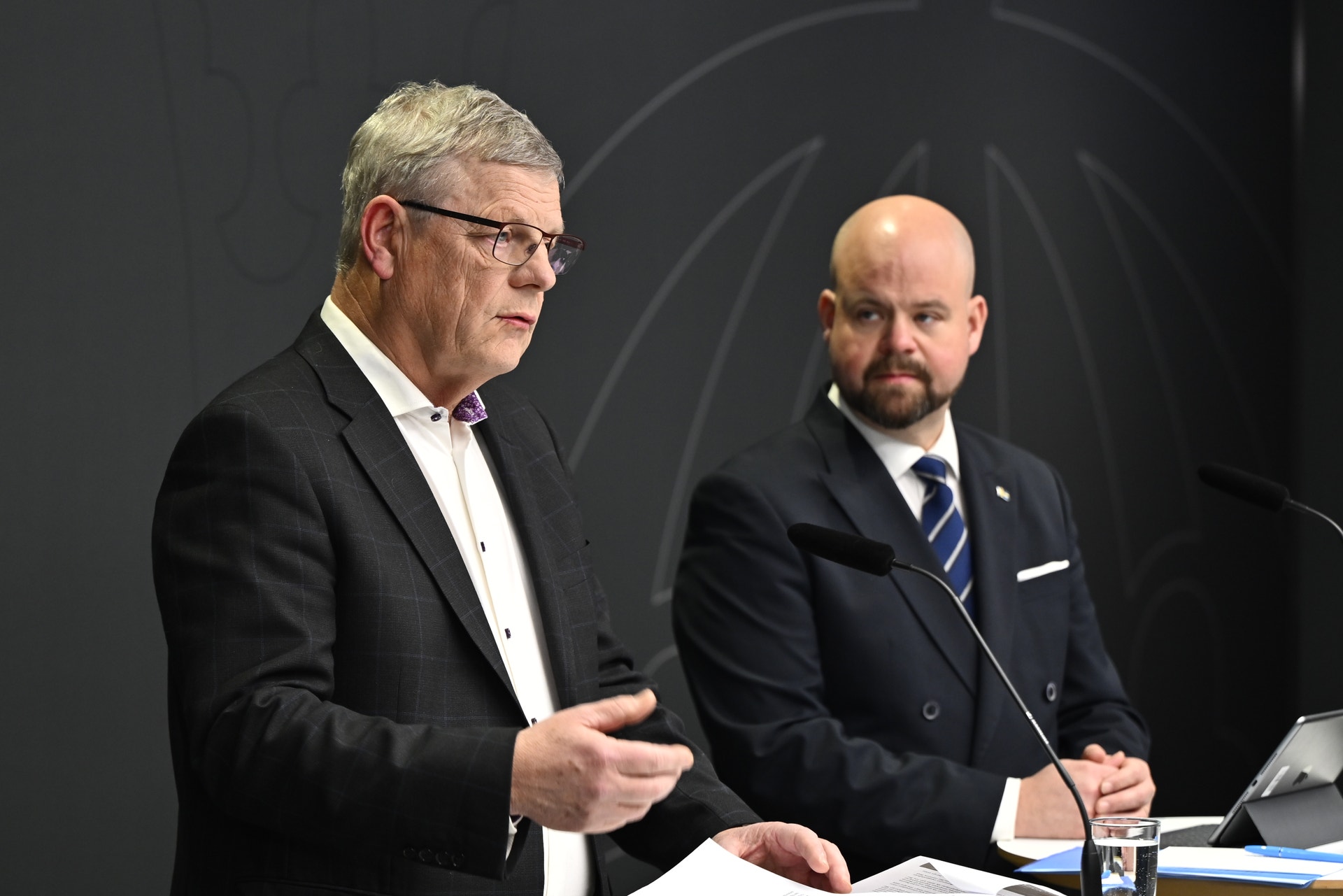 Två män i kostym presenterar på en scen. Mannen till vänster talar medan mannen till höger lyssnar.
