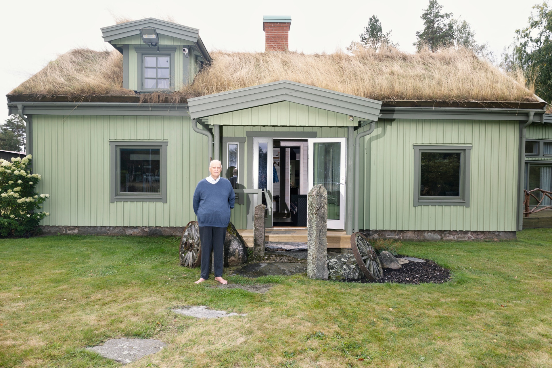 En person står framför ett grönt trähus med taktäckning av torv. Huset har klassisk nordisk arkitektur med ett litet fönster på taket och vita dörrar.