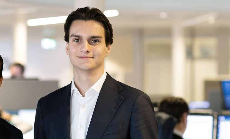 Porträttfoto av en man i svart kostym och vit skjorta, fotograferad i en modern kontorsmiljö med suddig bakgrund.