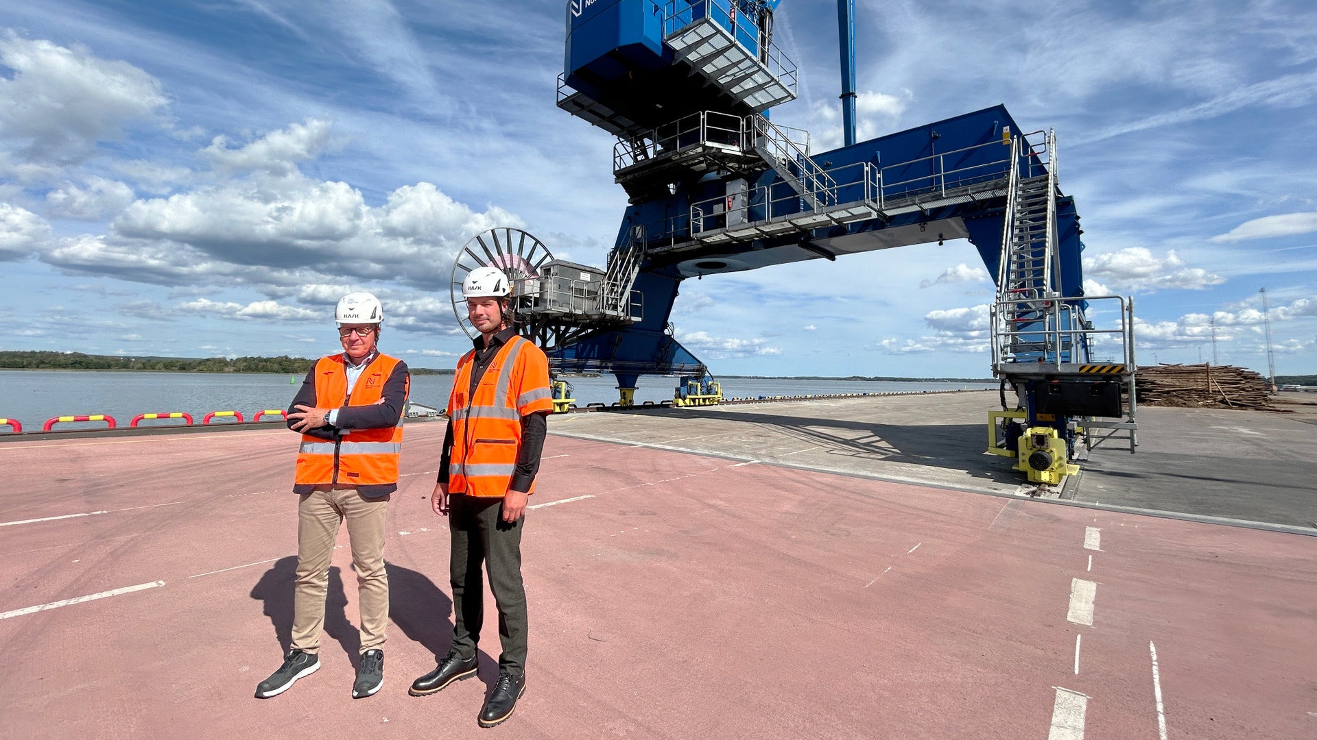 Två arbetare i orange säkerhetsvästor och vita hjälmar står vid en hamn framför en stor blå lastningsmaskin.