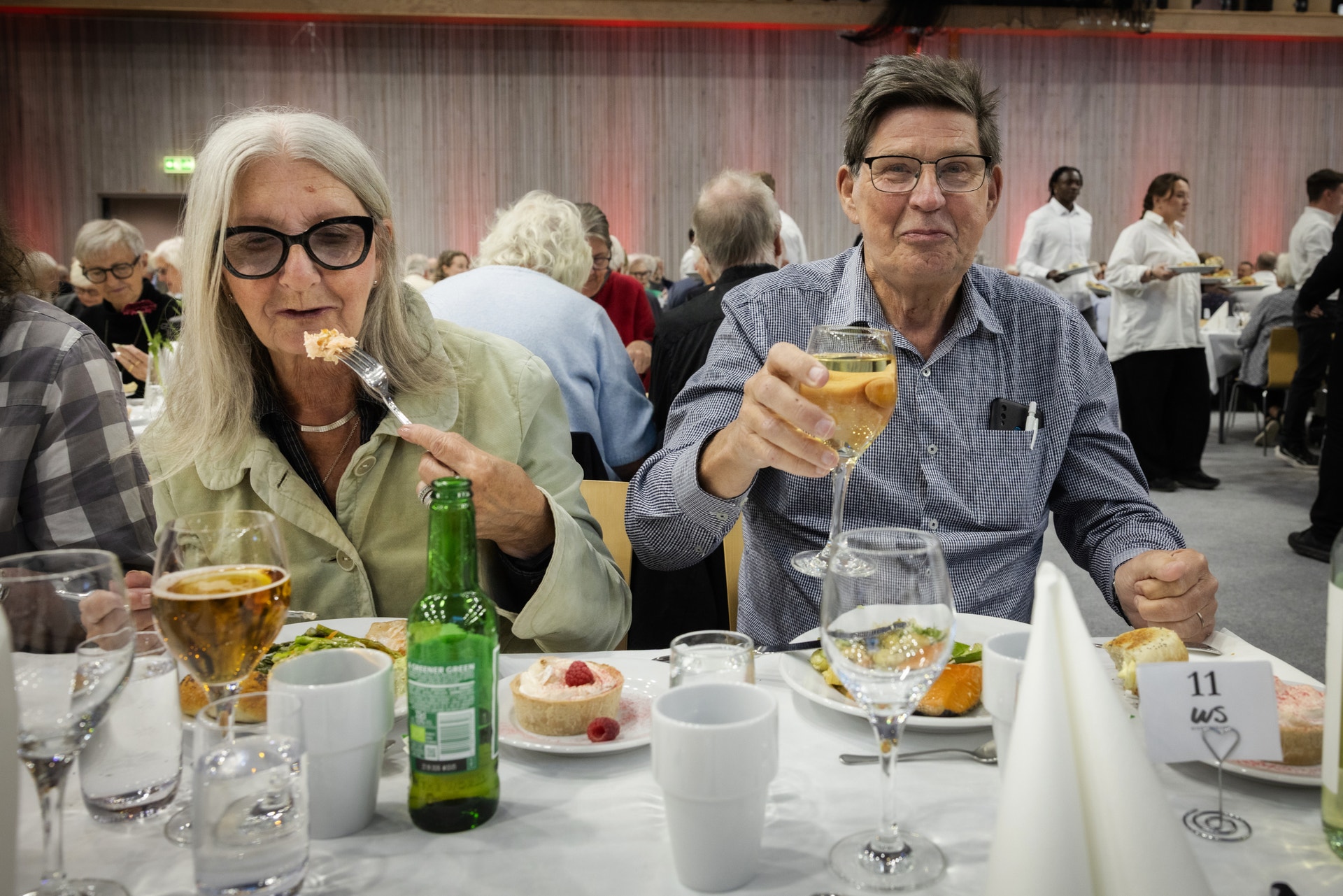 En man och en kvinna skålar med glas vid ett festbord. Flera andra gäster sitter i bakgrunden under ett event.